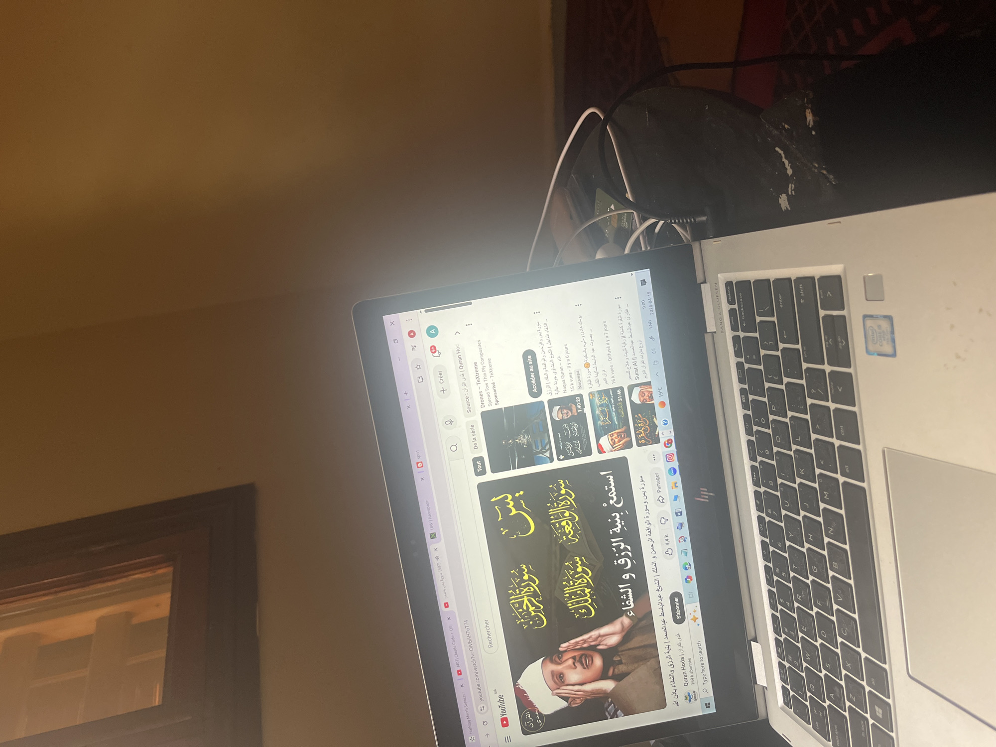 The image shows a laptop on a desk, displaying a YouTube video of Quranic recitations. The video features Arabic script and images of a reciter. The laptop's keyboard is visible, and headphones are plugged in nearby. The background includes a wall, a window frame, and a section of what appears to be a patterned surface. The video title displayed on the screen is "Listen with the intention of Rizk (sustenance) and healing." The image appears to have been taken indoors. The OCR text hints at location of Morocco, since "MA" is at the bottom left. No evidence indicates the specific city of Ouaouizert.
