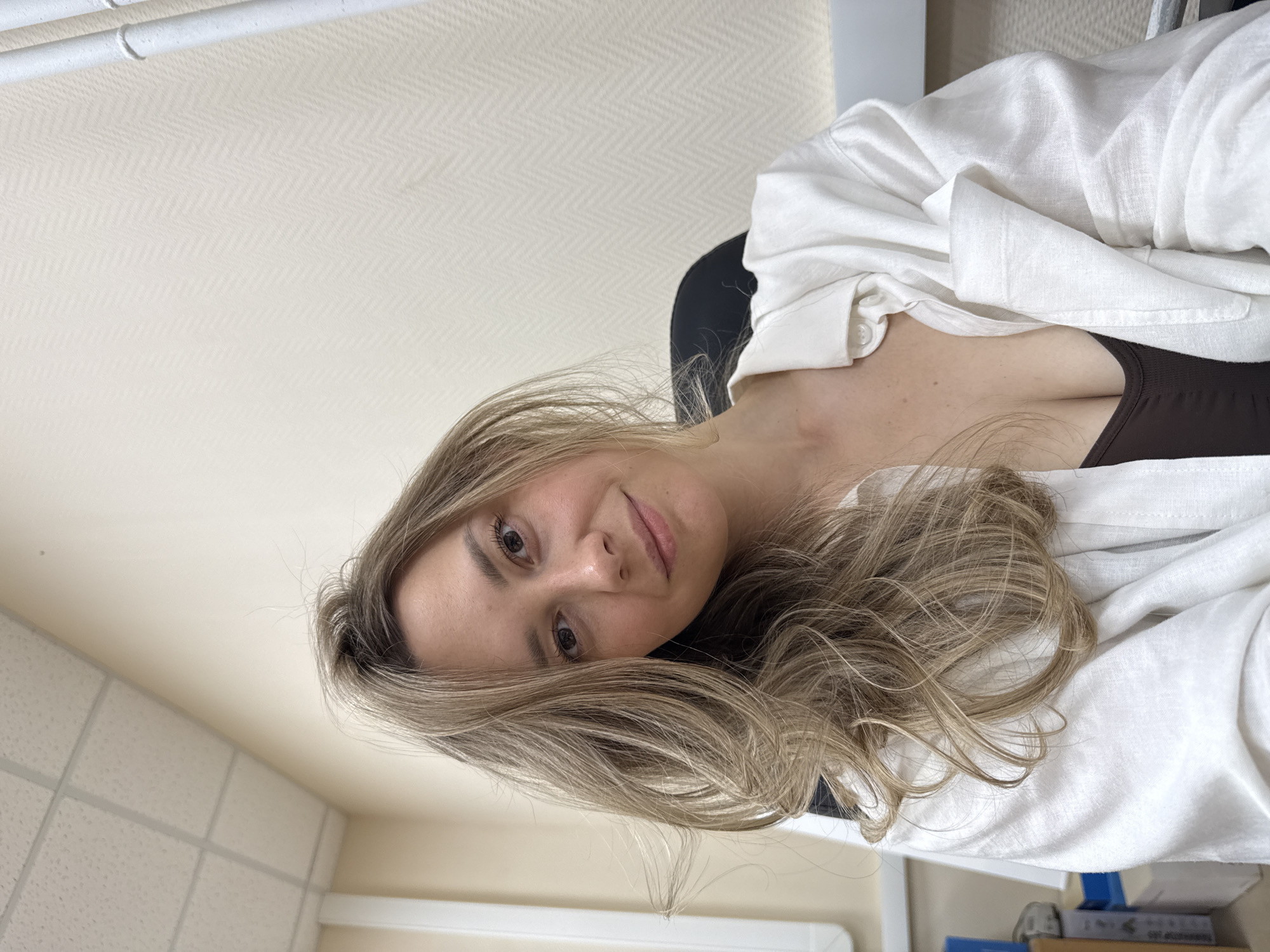 A woman with blonde hair and wearing a white shirt is seated. The location appears to be an interior room, possibly in an office or residential setting, judging by the type of ceiling tiles and wall paint. The woman is likely in Chaikovsky, Russia based on the prompt.