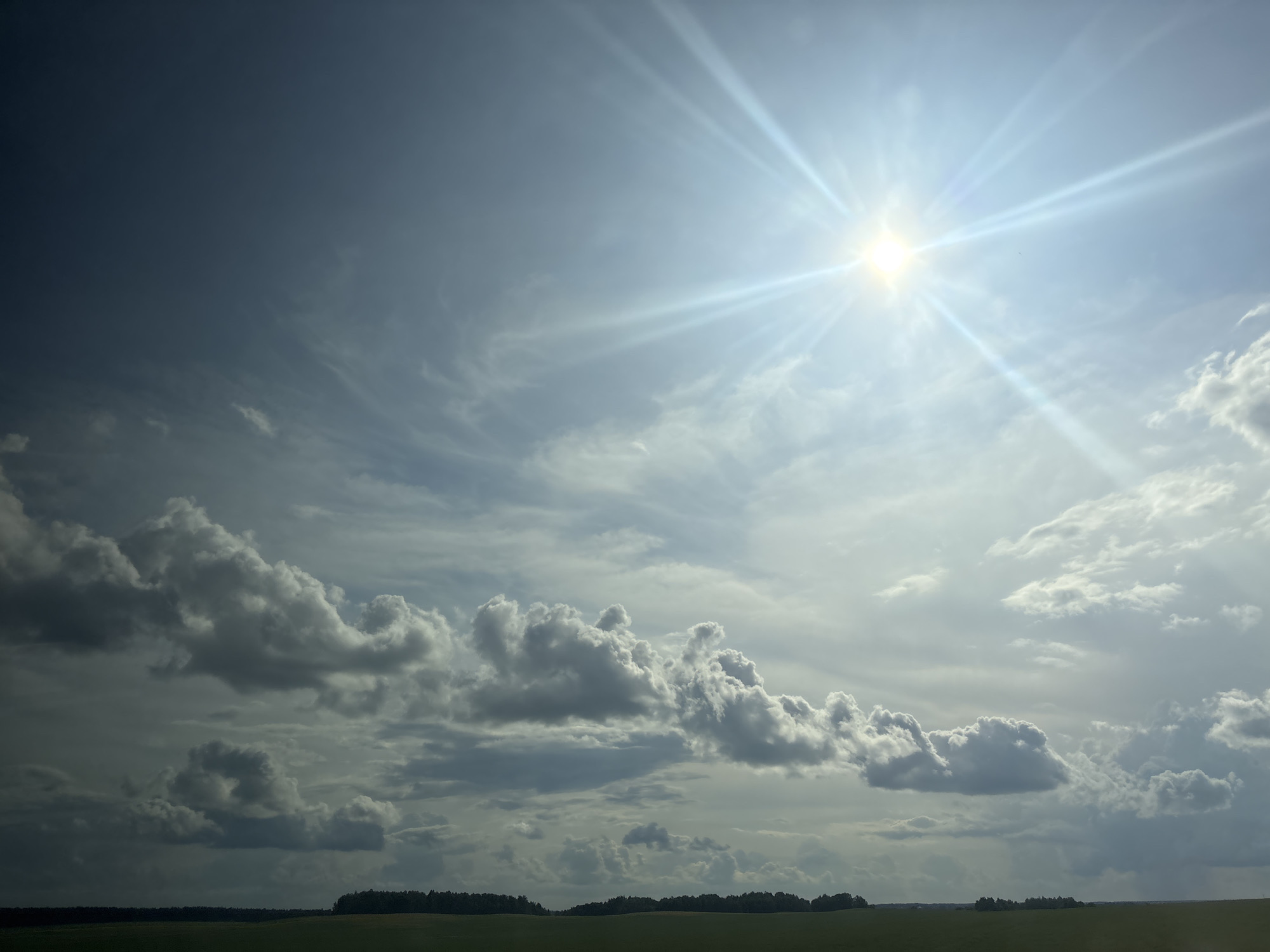 A wide-angle, landscape photograph captures a dramatic sky over a rural area. The sun, a bright, starburst-like object, is positioned in the upper right quadrant, casting brilliant rays across the hazy, blue sky. A mixture of dark, heavy clouds and lighter, wispy formations are scattered throughout, creating a dynamic contrast. The lower third of the image displays a dark, tree-lined horizon above a muted green field, suggesting an undeveloped, agricultural setting within the Сеніцкі сельскі Савет region of Belarus. No people or specific objects are identifiable in the scene.