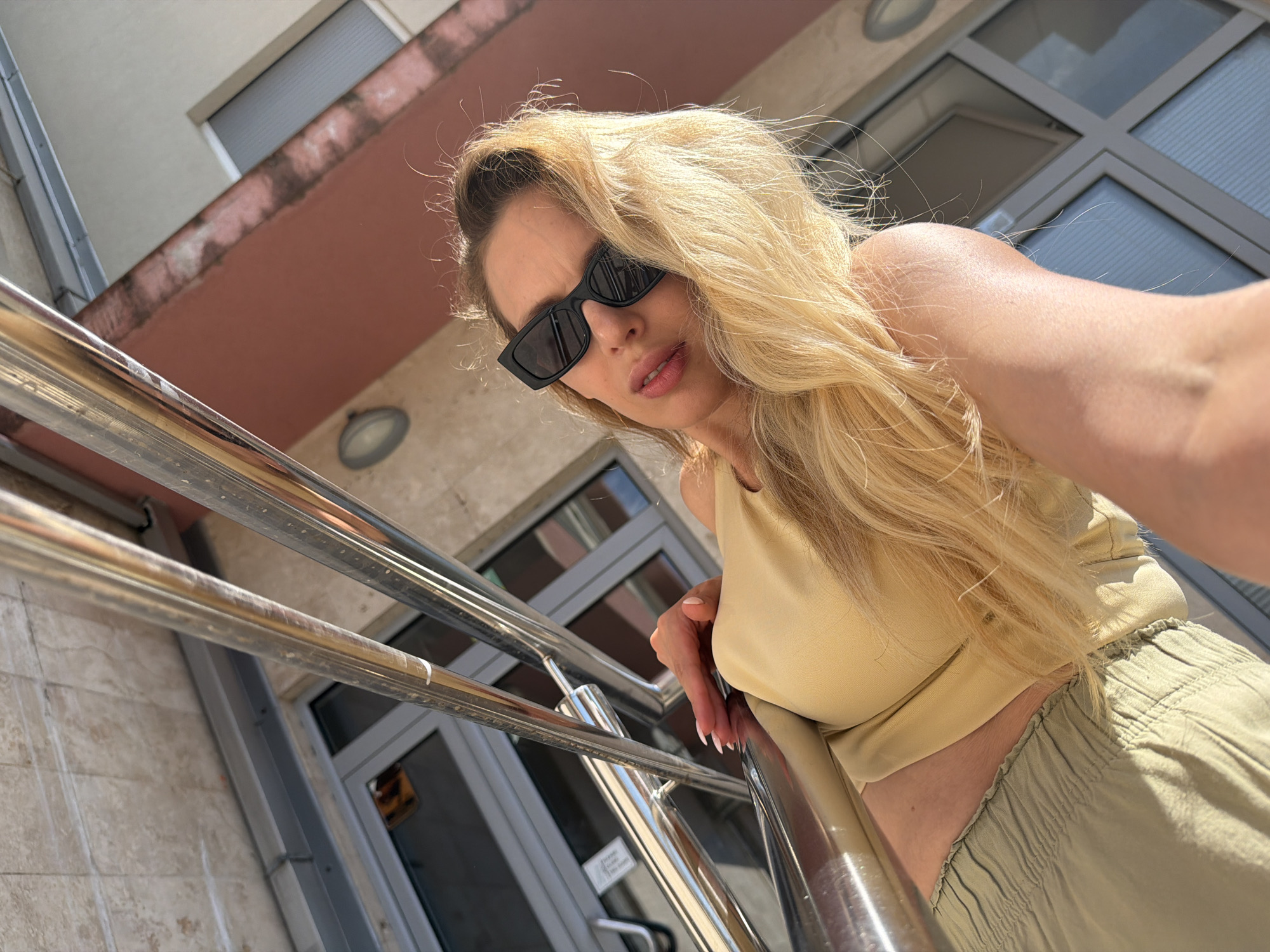 A blonde woman wearing sunglasses and a light yellow top stands outdoors, leaning on a metal railing. She is wearing light green pants. In the background, there is a building with windows and a reddish-brown facade. The scene appears to be in a city, possibly Budva, Montenegro, given the architectural style.