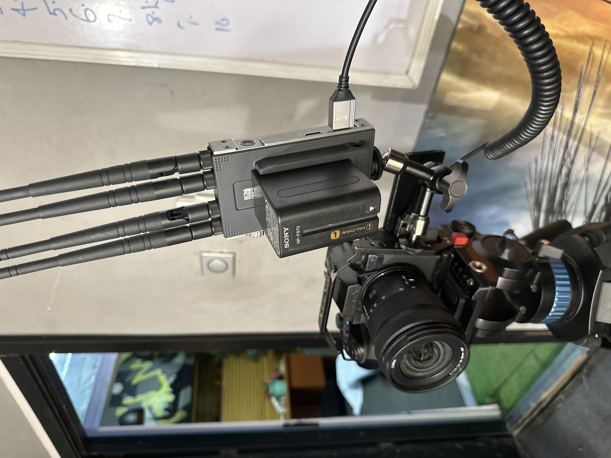 The image captures a detailed view of a professional video camera setup, likely in a studio or production environment in Ikeja, Nigeria. The primary subject is a mirrorless camera, specifically a Panasonic Lumix, equipped with a Panasonic Lumix S 20-60mm f/3.5-5.6 lens, visible from a downward angle. The camera is enclosed within a protective cage or rig, with various mounting points and accessories attached.
Mounted above the camera is a wireless video transmitter/receiver unit, distinguishable by its gray metallic housing and three prominent external antennas. A Sony NP-F970 InfoLITHIUM L battery powers this unit, directly attached to its side. A black USB cable is plugged into the top of the wireless unit, connecting it to an unidentifiable device. Another coiled black cable extends from the camera rig upwards, suggesting a power or video connection.
In the background, a white whiteboard is visible with handwritten numbers "1 2 3 4 5 6 7 8 9 10" in blue marker. Reflections on a surface below the camera reveal a blurred indoor scene, including glimpses of furniture and possibly other objects. The setup indicates active use or preparation for video recording or live production, utilizing wireless video transmission for monitoring.