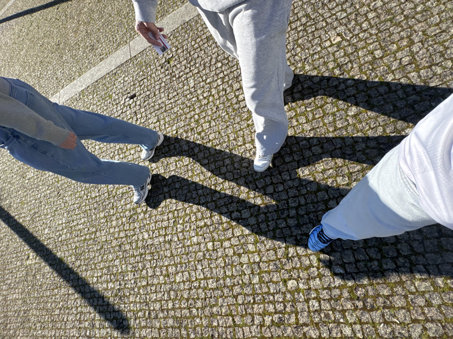 Three individuals are observed on a cobblestone pavement characteristic of Portuguese cities, likely in Bragança, Portugal. The ground is composed of small grey stone blocks, with visible green moss in the interstices. A faint white line traverses the pavement. Strong, elongated shadows are cast on the ground, indicating direct sunlight from a low angle.
The first individual, positioned towards the upper left, is seen from the mid-thigh down, wearing light blue, loose-fitting jeans and white athletic shoes with dark accents. A grey garment is visible above the waistline.
The second individual, located towards the mid-right, is also seen from the mid-thigh down, dressed in light grey sweatpants and white athletic shoes. This person holds a white smartphone with a purple-patterned screen in their right hand, oriented away from the viewer.
The third individual, whose lower legs and feet are visible at the bottom-left, appears to be the photographer. This person wears light-colored (possibly white or light blue) pants and blue athletic shoes with black detailing.
The individuals are in close proximity, standing or walking on the sunlit pavement. The specific location within Bragança cannot be precisely identified from the image content beyond being a public outdoor area with traditional Portuguese pavement.