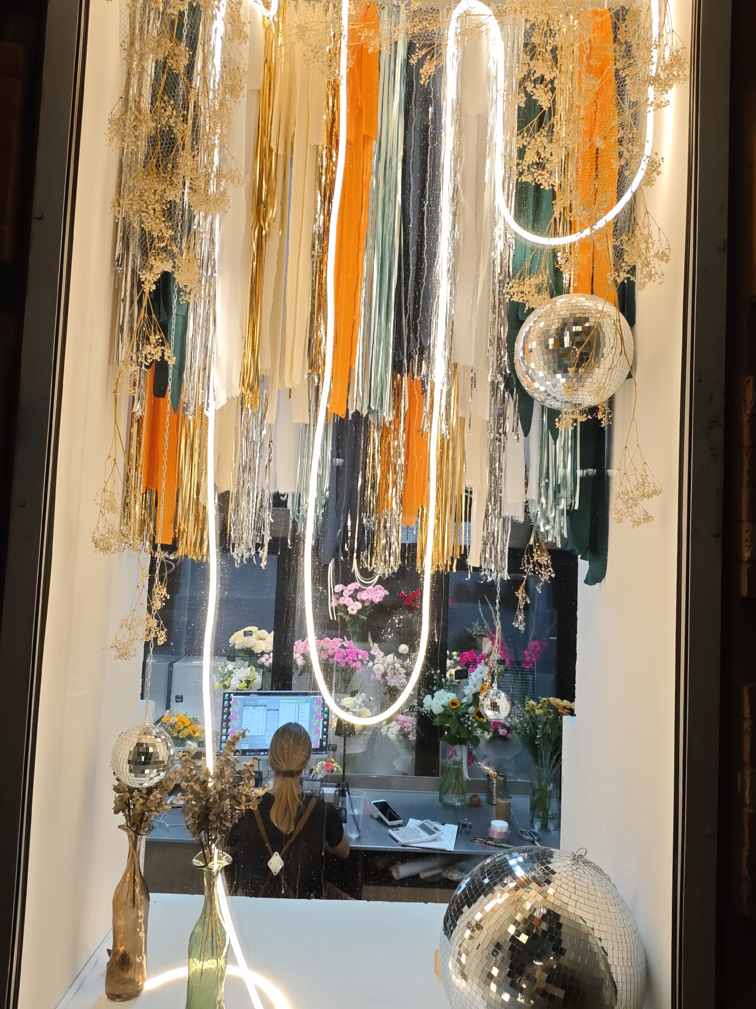 In the city of Unknown, Unknown, a florist shop window is decorated with metallic streamers and dried flowers hanging from the ceiling. Two disco balls are visible, one larger one on the right and a smaller one hanging near the top. Inside the shop, a woman with blonde hair pulled back in a ponytail, wearing a black t-shirt and a white apron, is seated at a counter in front of a computer monitor. She is facing away from the camera and appears to be working. On the counter, there are various floral arrangements in vases, as well as other items like scissors, a calculator, and a credit card reader. The overall scene suggests a festive and possibly artistic display within a retail environment.