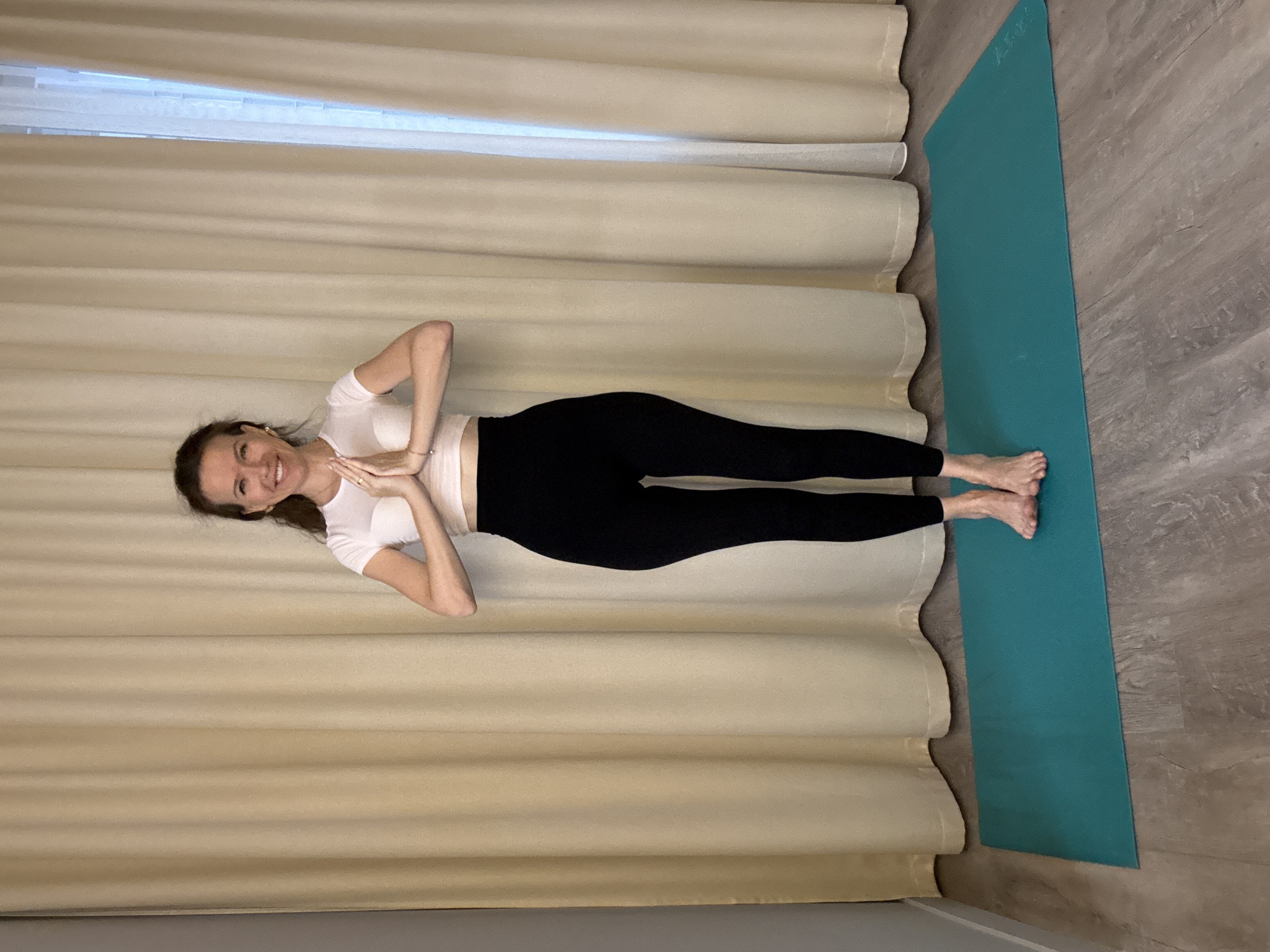 A woman with brown hair is standing on a teal yoga mat on a wooden floor in front of a beige curtain. She is wearing a white crop top and black leggings. Her hands are pressed together in front of her chest in a prayer pose. She is smiling and looking at the camera. The scene appears to be indoors, possibly a home or yoga studio, in Dolgoprudny, Russia.