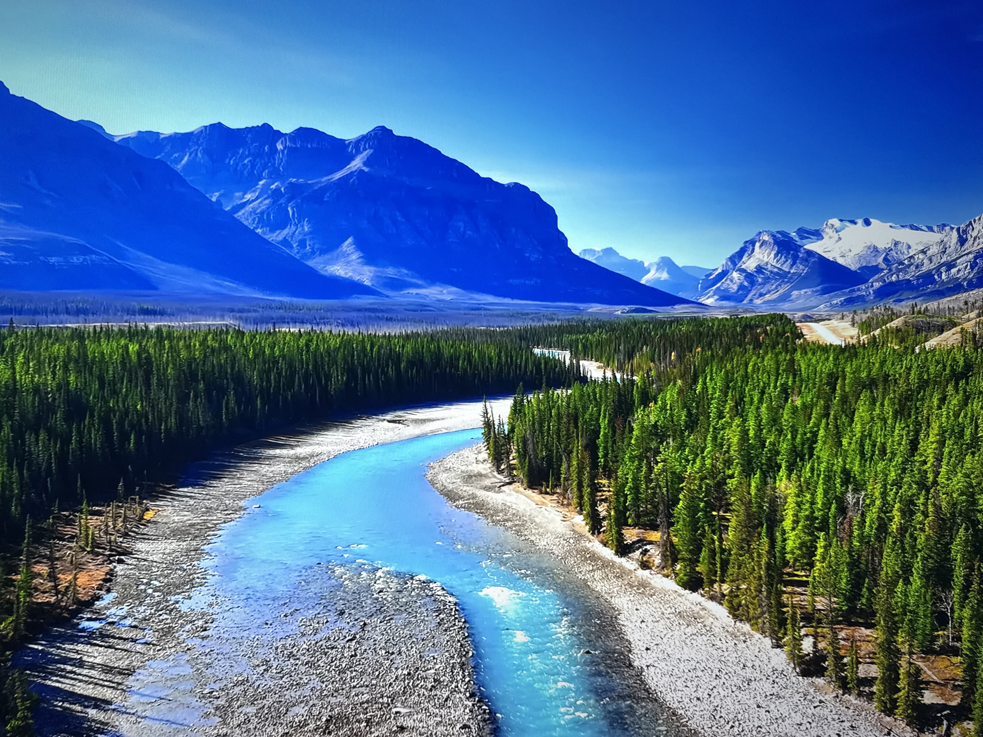 This image depicts a natural landscape, likely a river valley in a mountainous region. A vibrant blue river snakes through the foreground, bordered by a rocky shore on one side and a dense evergreen forest on the other. The forest extends to the midground, where it meets a vast, flat expanse. In the background, towering, jagged blue mountains dominate the horizon, their peaks sharp against a clear, bright blue sky. The overall scene is one of wilderness and natural beauty, with strong contrasts between the colors of the water, the trees, and the mountains. There is no indication of any urban environment or the city of Burgas, Bulgaria, in this image.