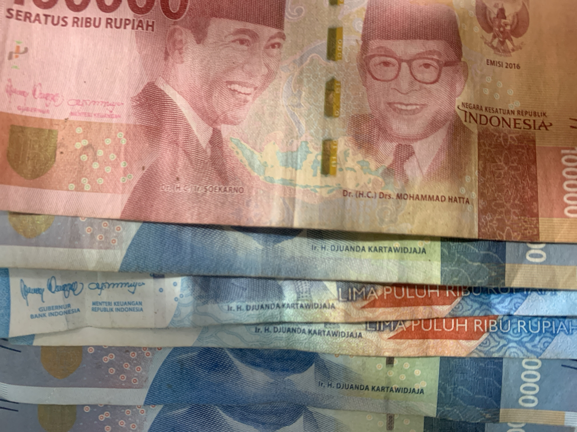 This media file displays a close-up, high-angle view of stacked Indonesian rupiah banknotes. The primary subjects are the portraits of Indonesian historical figures featured on the currency. On the top banknote, a red one, two men are visible: Soekarno on the left and Mohammad Hatta on the right. The text "SERATUS RIBU RUPIAH" indicates the denomination of 100,000 Rupiah. Below the portraits, the names "Dr. H.C. Ir. SOEKARNO" and "Dr. (H.C.) Drs. MOHAMMAD HATTA" are visible. The emission year "EMISI 2016" is also present, along with "NEGARA KESATUAN REPUBLIK INDONESIA".
Below the top banknote, several other denominations are partially visible. These include banknotes with blue and green color schemes. Textual elements on these lower banknotes include "Ir. H. DJUANDA KARTAWIDJAJA", "GUBERNUR BANK INDONESIA", and "MENTERI KEUANGAN REPUBLIK INDONESIA". The phrase "LIMA PULUH RIBU RUPIAH" (Fifty Thousand Rupiah) is also clearly legible on some of these.
The lighting suggests an indoor setting or possibly an overcast day, as there are no strong shadows. The focus is on the details of the banknotes, highlighting their design and text. The image's composition, with the overlapping bills, creates a sense of abundance or a collection of money. There are no overt emotional cues, but the image is informative in its display of currency. The specific location mentioned as Sungai Meriam, Indonesia, provides a geographical context for the currency being Indonesian.