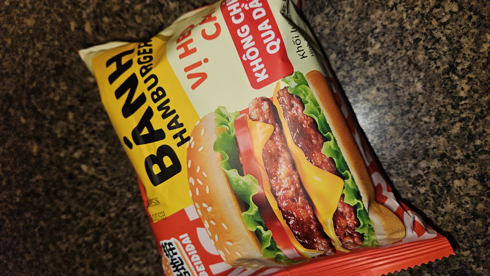 The image shows a close-up of a package of "Bánh Hamburger" flavored crackers. The package is predominantly yellow, red, and white, with a picture of a cheeseburger. Text in Vietnamese and Chinese is visible on the packaging. The background is a dark, speckled surface, possibly a countertop. The crackers are most likely in Cam Duc, Vietnam.