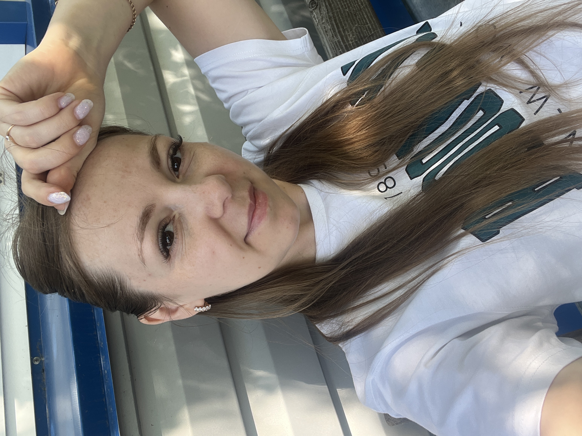 A selfie taken outdoors shows a Caucasian woman with long brown hair and fair skin. She is wearing a white t-shirt with a green graphic print and a ring on her right hand. Her left hand is resting on her forehead, and she is wearing small hoop earrings with rhinestones. The background is a light-colored, corrugated wall with a blue stripe at the top, suggesting an outdoor structure or building. The lighting indicates it is daytime, and there are shadows cast on the wall. The woman is smiling slightly, and her gaze is directed at the camera. Based on the provided information, the image was taken in the city of Shadrinsk, Russia.