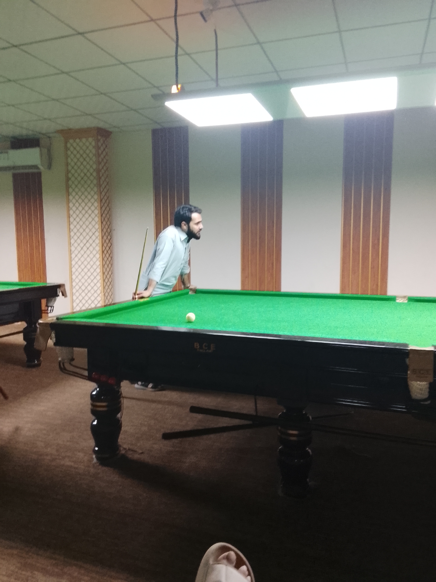 The media file captures an indoor scene in what appears to be a snooker or billiards hall. The main subject is a man with a beard, wearing a light-colored long-sleeved shirt, leaning over a snooker table. He is holding a snooker cue, positioned as if about to take a shot, and is looking intently at a white snooker ball on the green felt surface of the table.
In the foreground, the edge of another snooker table is visible on the left, and partially visible bare feet, possibly belonging to the photographer, are seen at the bottom right. The table in the foreground features ornate dark wooden legs and has the text "B.C.F." inscribed on its side.
The setting includes walls decorated with vertical wooden panels, some featuring a lattice pattern. An air conditioning unit is mounted on the left wall in the background. The ceiling is tiled with multiple bright, rectangular fluorescent light panels providing ample illumination. A dark wire descends from the ceiling above the man. The floor is carpeted in a dark brown or maroon color. The man appears focused on his game. The artificial lighting suggests no specific time of day.