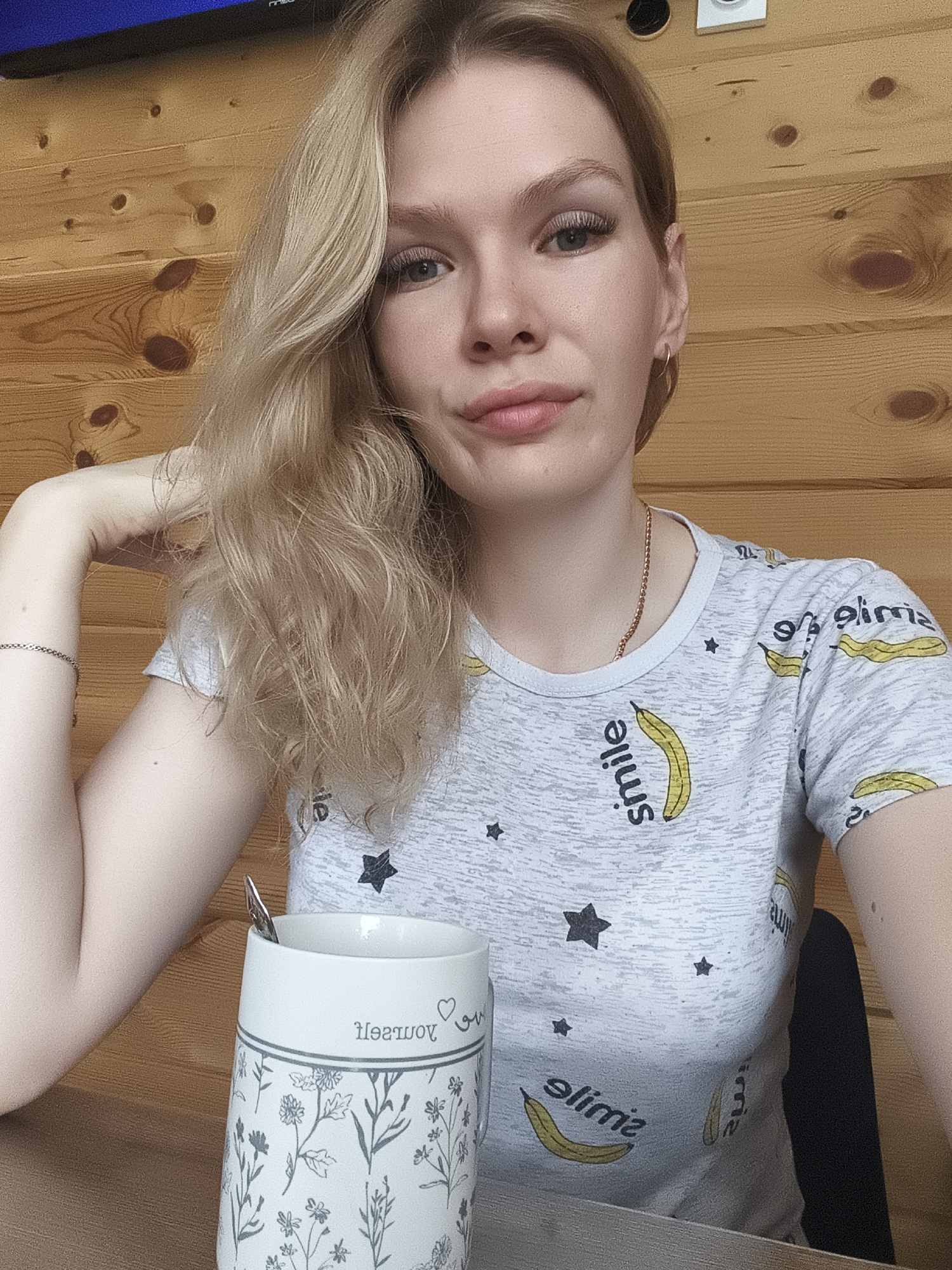 A young woman with blonde hair sits at a wooden table, holding a white mug with a floral design and a spoon. She is wearing a gray t-shirt with a pattern of bananas and the word "smile." The background is a wooden wall. The image appears to be taken indoors, likely in a home or cafe. There is no visible indication of the city of Русский Вожой, Russia, within the image itself.