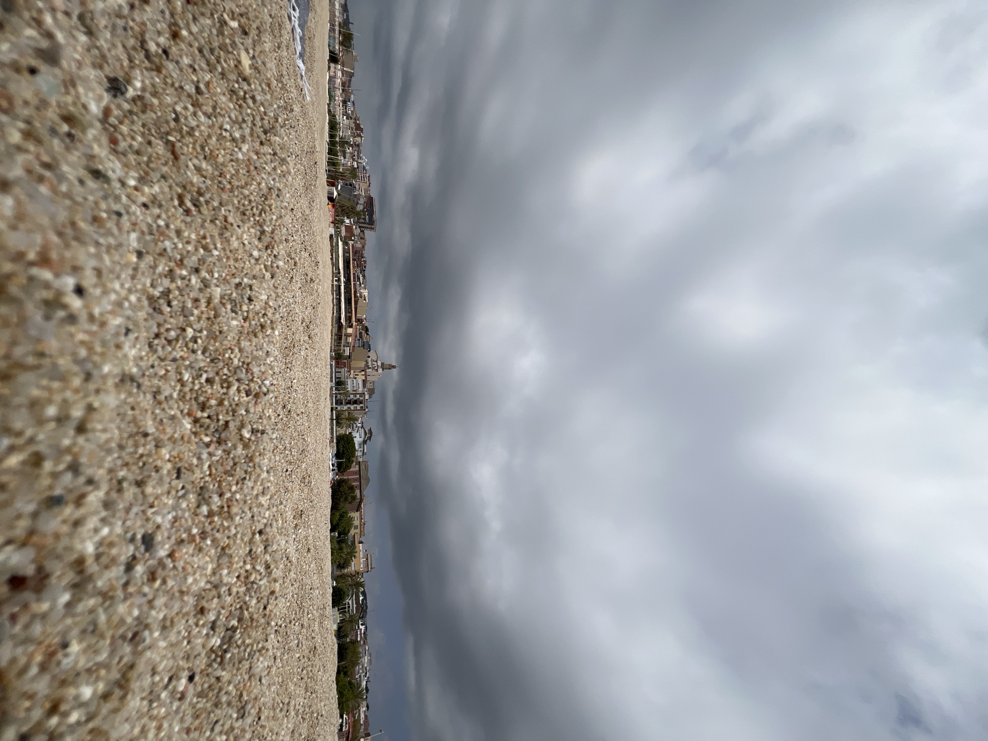 The image depicts a close-up view of a pebble beach in El Masnou, Spain. The foreground is dominated by the granular texture of the pebbles, with some appearing wet from the receding tide. In the mid-ground, the coastline of El Masnou is visible, featuring a cluster of multi-story buildings, hotels, and shops. Distinctive architectural elements include a prominent church tower with a clock face, as well as balconies and rooftop terraces on the residential and commercial structures. Palm trees and other vegetation are interspersed among the buildings. The background is filled with a heavy, overcast sky, characterized by dark, dramatic storm clouds that obscure the horizon. The overall atmosphere is somber and foreboding, suggesting an impending storm or recent rainfall. There are no discernible people or active events captured in the scene.