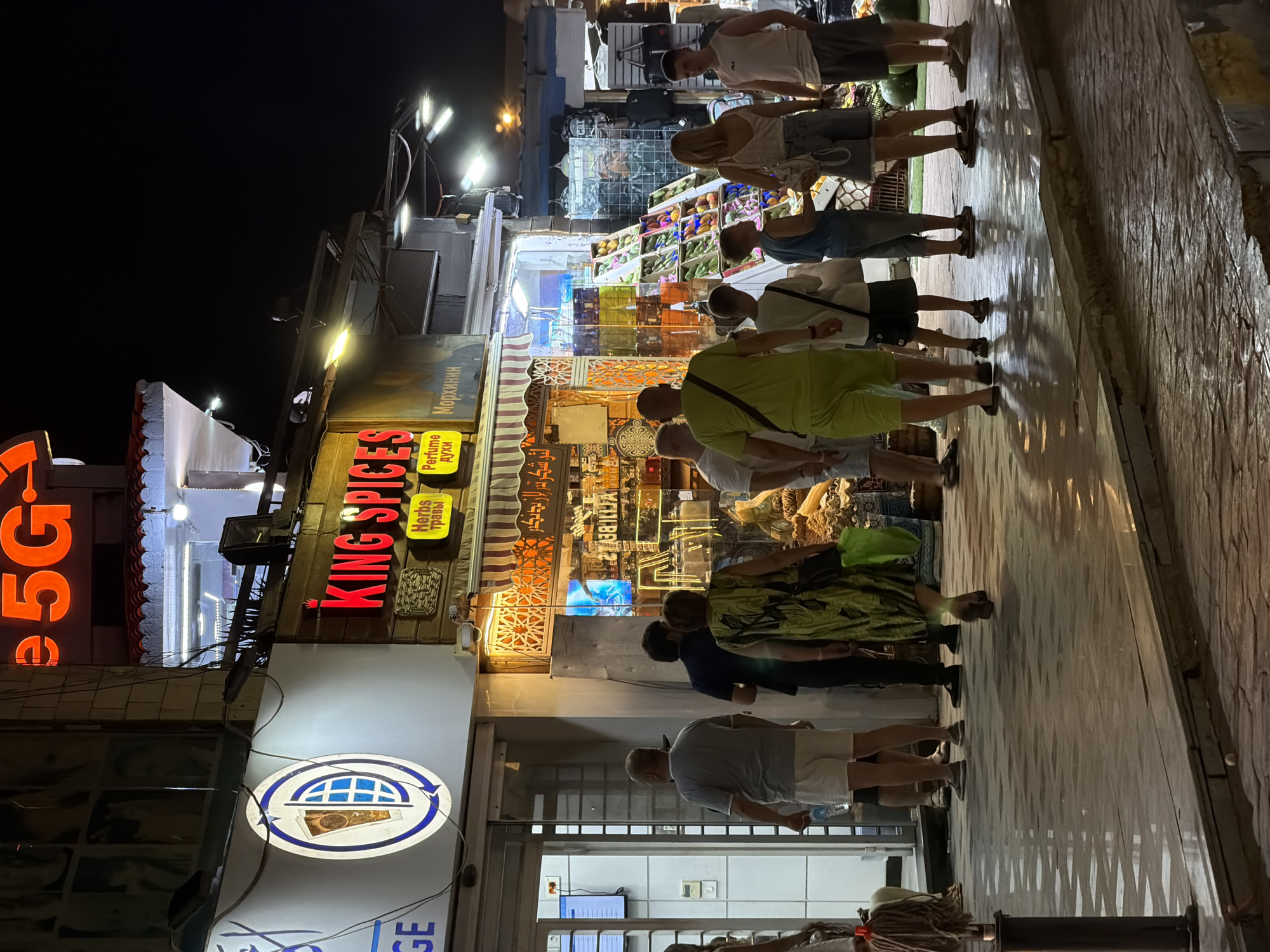 The image depicts a nighttime street scene in Sharm El Sheikh, Egypt, specifically in a commercial area with illuminated shops. A prominent sign reads "KING SPICES" in red lettering, with smaller yellow signs below indicating "Herbs" and "Perfume" in Cyrillic and English. The shops appear to sell various goods, including spices, perfumes, and fruits displayed in crates. Several people are walking on the paved sidewalk, which reflects the ambient light. The architecture suggests a tourist-oriented location with awnings and decorative elements.