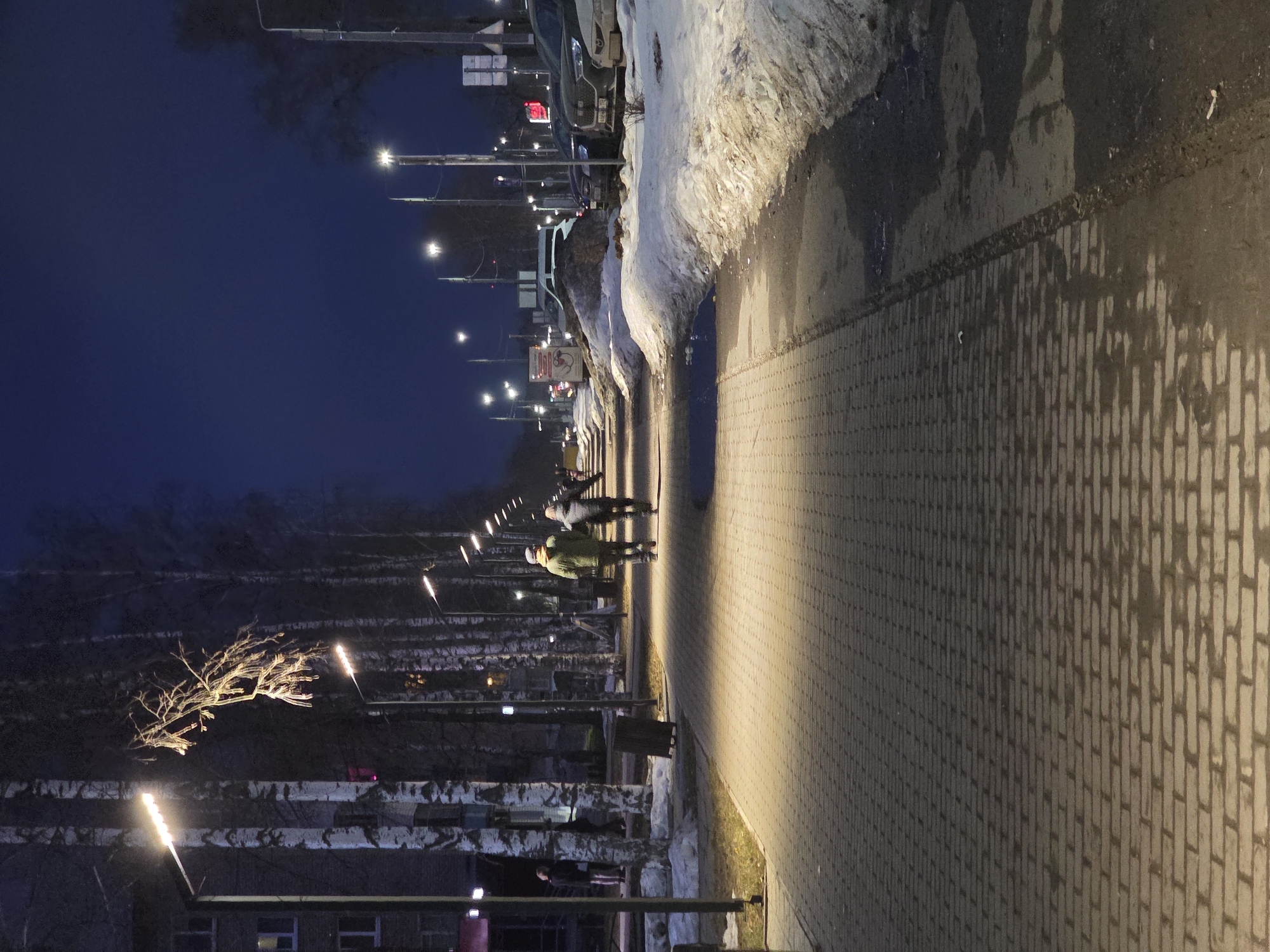 The image depicts a sidewalk at night in what appears to be a snow-affected environment, likely near a street. The scene is in the city of Chaikovsky, Russia. Streetlights illuminate the path, and snow piles up along the roadside. Several individuals can be seen walking. There are bare trees along the left side. There is a building shown on the lower-left corner with at least one window showing. The sidewalk appears to be paved with rectangular stone blocks.