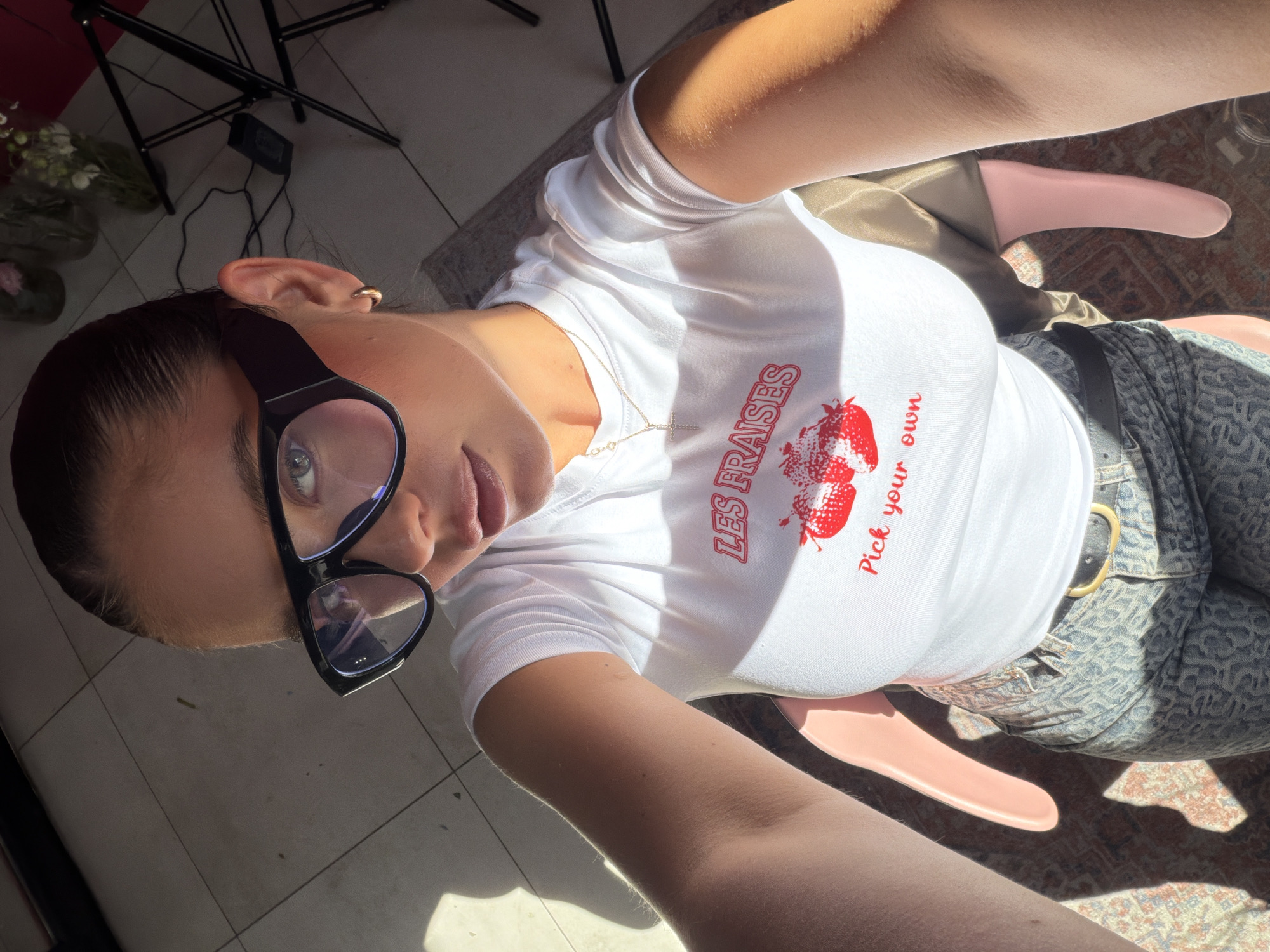 A woman with dark hair pulled back is wearing black glasses, a white t-shirt with red text and strawberry graphics, and patterned jeans with a brown belt. She is sitting on a pink chair, indoors with sunlight streaming in, illuminating a tiled floor and an area rug. Tripod and vases of flowers are visible in the background. The photo appears to be a selfie taken from above. There is no indication of the city of Krasnoyarsk, Russia in this image.