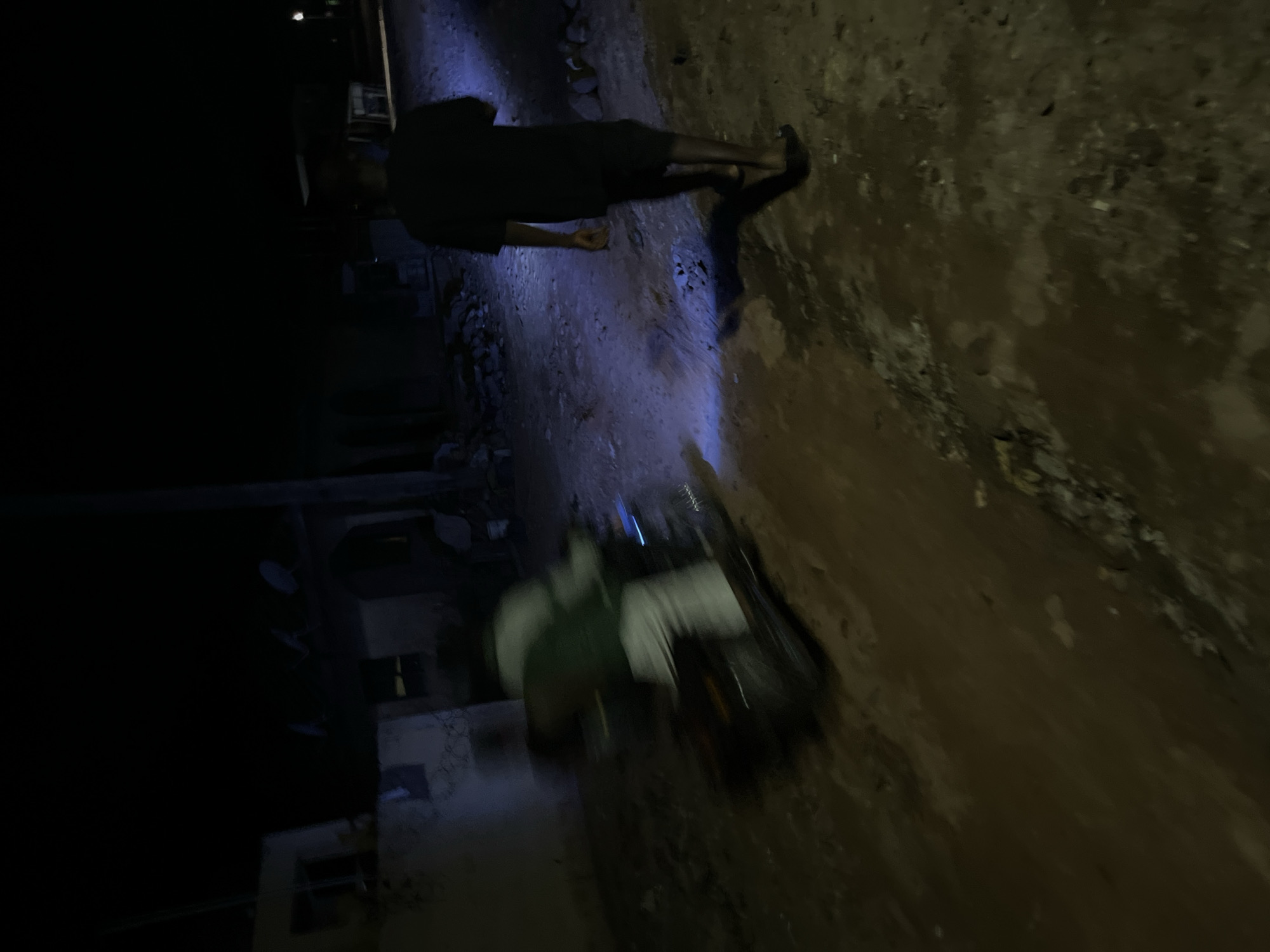In the city of Unknown, Unknown, at night, a person walks away from the camera on a dirt road, illuminated by a bright blue light. The person is wearing a dark t-shirt and shorts, and their shadow is cast behind them. To the left of the frame, a motorcycle is seen in motion, with a blurred figure on it wearing a green shirt and white pants. The background consists of dark buildings and debris, typical of a poorly lit urban environment. The overall scene suggests a moment of nocturnal activity in an unpaved area of the city.
