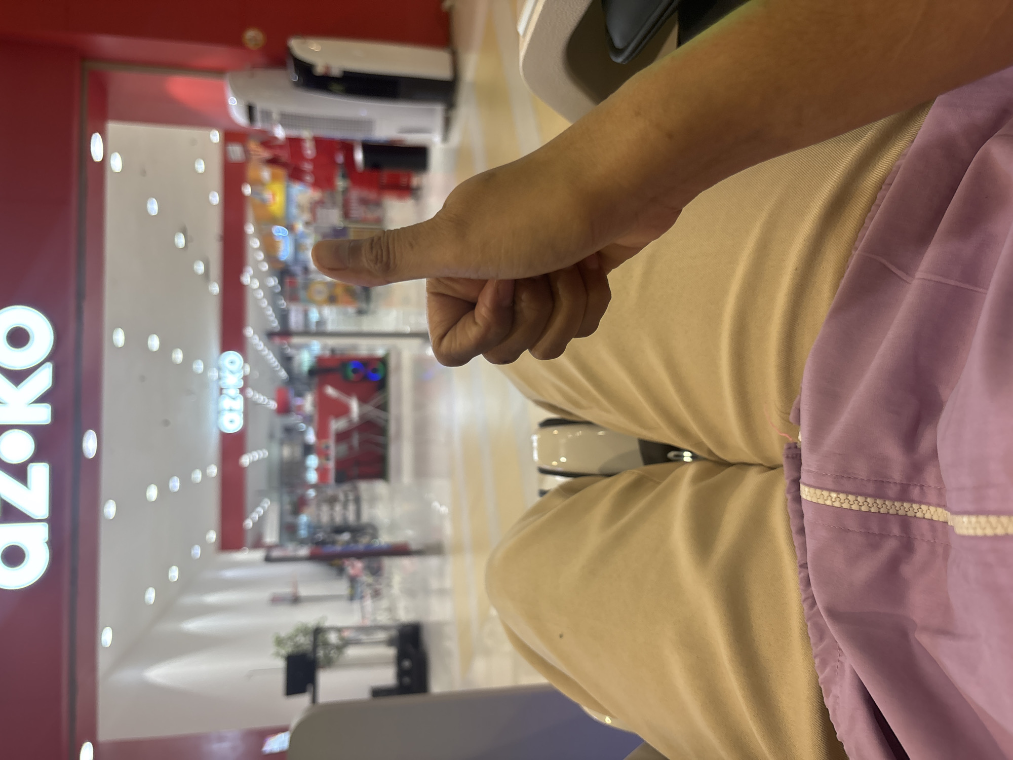 A first-person perspective shot shows a person's right hand giving a thumbs-up gesture while seated in what appears to be a shopping mall. The person is wearing light brown trousers and a lilac jacket. In the background, the sign for "AZKO" is visible above the entrance of a store, suggesting a retail environment within a city, possibly in North Jakarta, Indonesia. The store appears to sell electronics and household appliances, with displays of various products like fans and possibly computers visible. The floor of the mall is tiled with a pattern of yellow and white squares.
