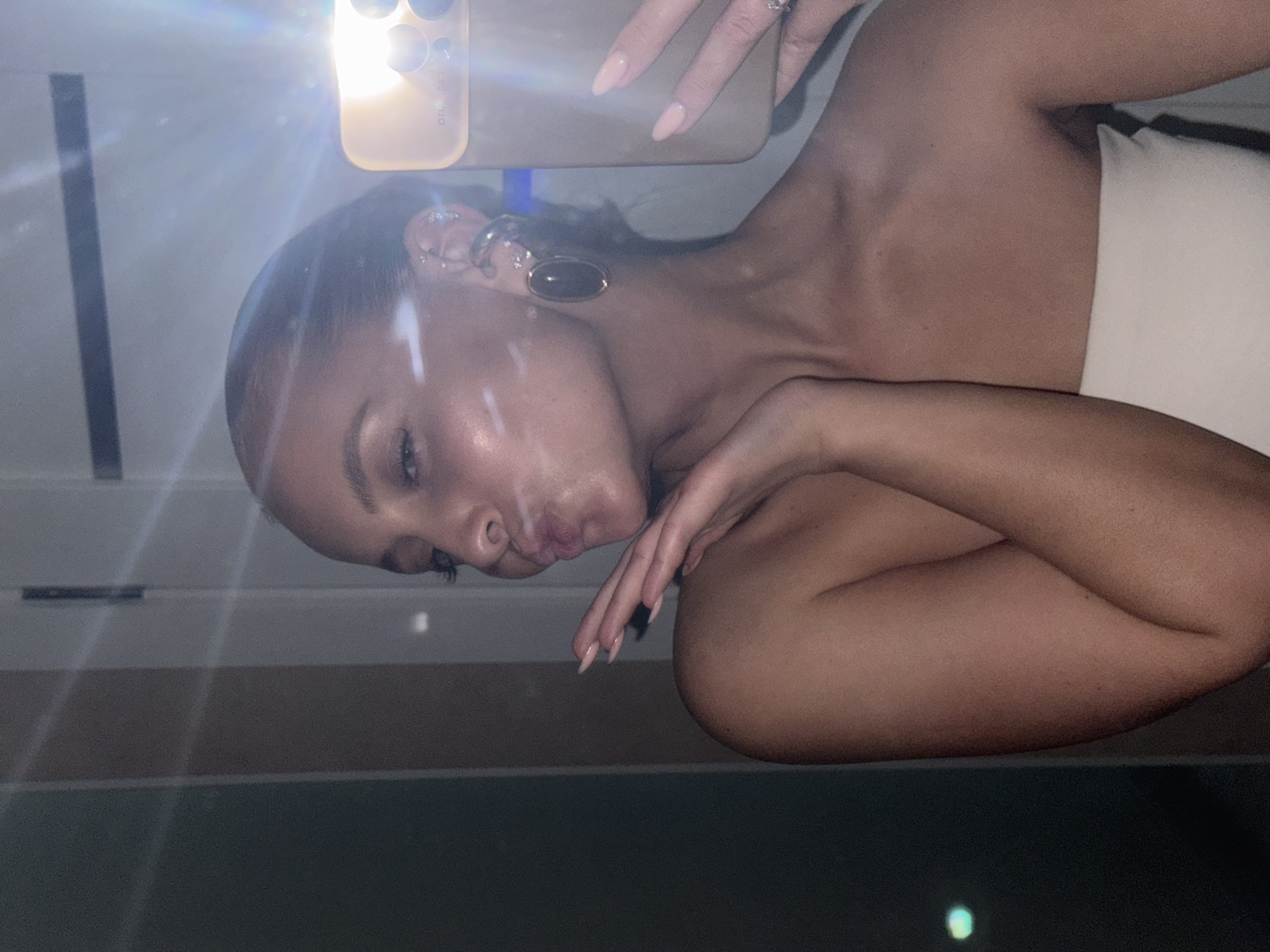A woman with tan skin is taking a selfie. She is pouting her lips and holding a phone with her right hand. She is wearing a strapless top or dress. Her left hand is near her face. She has several earrings in her left ear, including a large, dark-colored earring. The location is not discernible from the image; therefore, it is not possible to determine if it is Chaikovsky, Russia. The image is overexposed with a bright light reflection from the phone camera. The background is blurry, suggesting an indoor setting.
