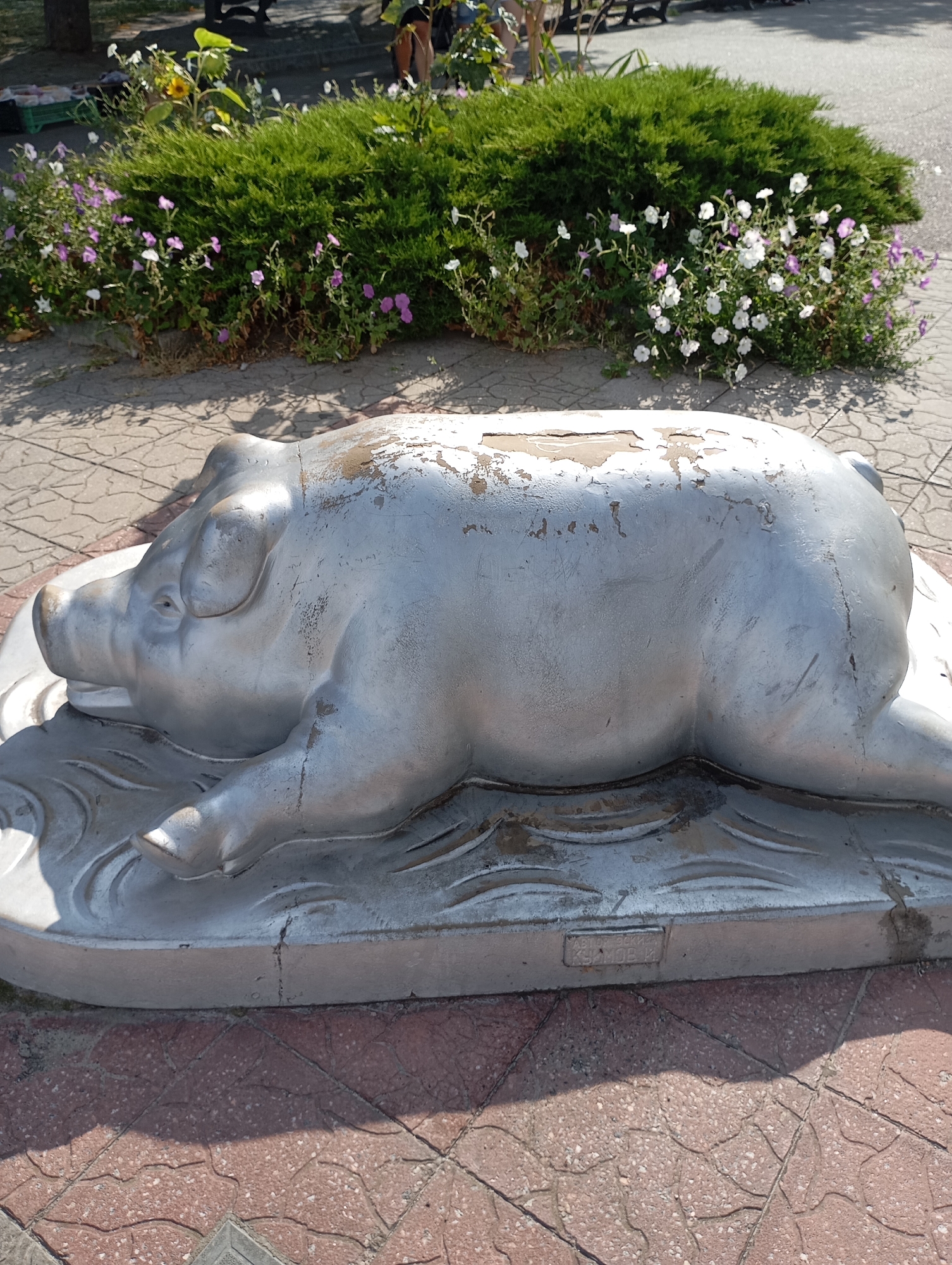 A silver statue of a pig lies on its side on a decorative base with wavy patterns. The statue is weathered, with peeling paint and scratches visible. It appears to be a public art installation in a park-like setting. Behind the pig statue, a dense green bush is in bloom with small white and purple flowers. The ground is paved with reddish-brown interlocking stones. In the background, blurred figures of people and trees are visible. A green plastic crate is also present in the background to the left. The scene is illuminated by bright sunlight, casting shadows. The statue is located in the city of Horishni Plavni, Ukraine, possibly within a public park or square.