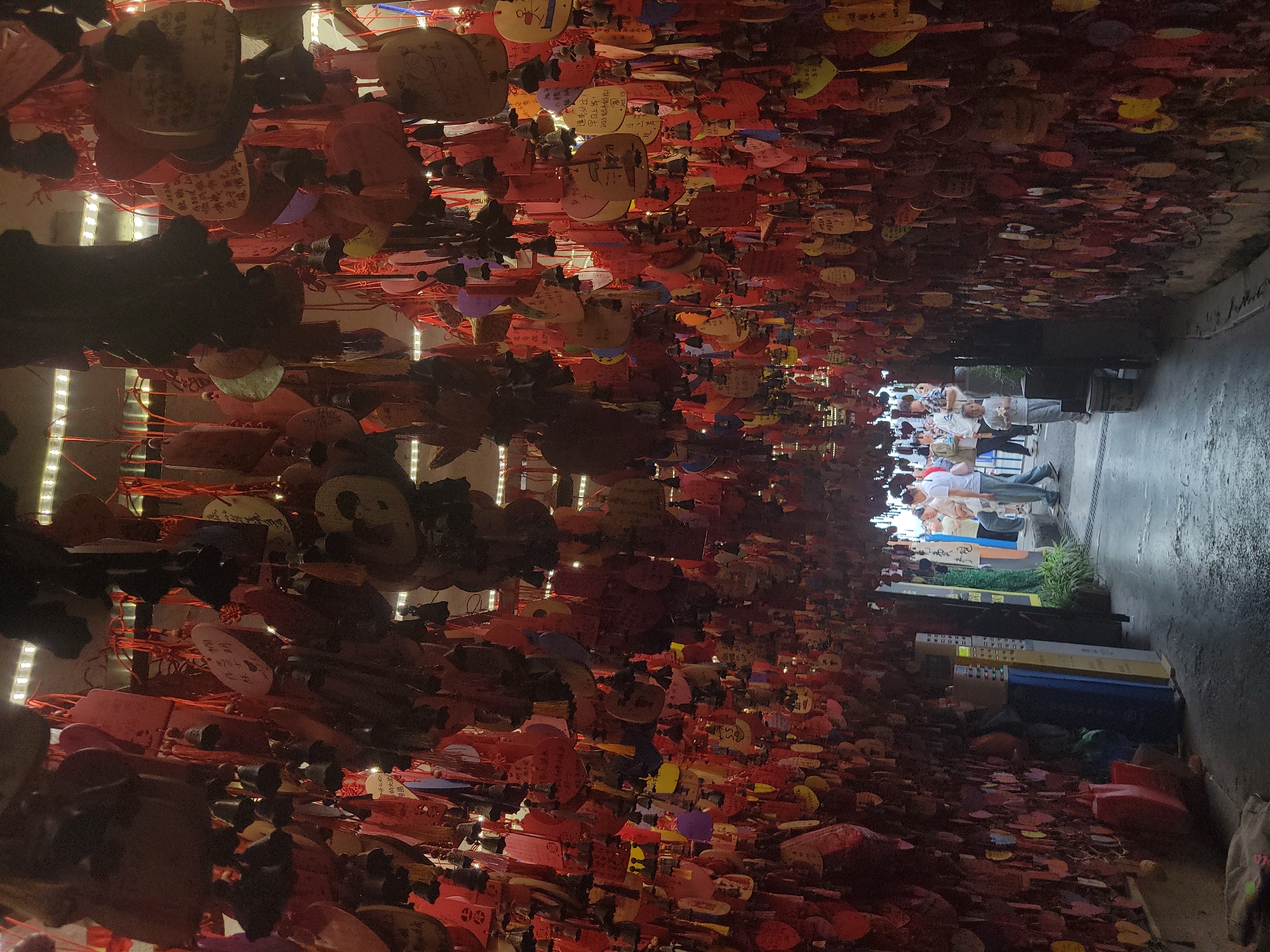 Here's a detailed description of the image:
**General Scene:**
The image presents a congested space covered with numerous hanging objects. These objects, primarily heart-shaped and circular, appear to be decorated with writing and tied with red strings. The scene suggests a wish-making or memorial site. There are people walking in the background, indicating an alley.
**Specific Elements:**
* **Hanging Objects:** The primary visual element is the dense collection of heart-shaped and circular objects hanging from above. They are brightly colored, including red, yellow, pink, and other hues. Many seem to have handwritten text, likely wishes or dedications.
* **Lighting:** The area is lit with parallel LED strip lights.
* **People:** In the background, a few people can be seen walking along a passageway.
* **Surroundings:** A section of a passageway is visible, partially illuminated by natural light. Vending machines and planters are visible along the alleyway, and a tiled floor is visible.
**Possible Location (based on visual cues and provided information):**
While it is difficult to be certain without further context, based on the provided text and visual cues, this location could be in Yuzhong District, China. The hanging objects and the overall environment suggest a tourist area or a place with cultural significance where people commonly make wishes or leave memorials.