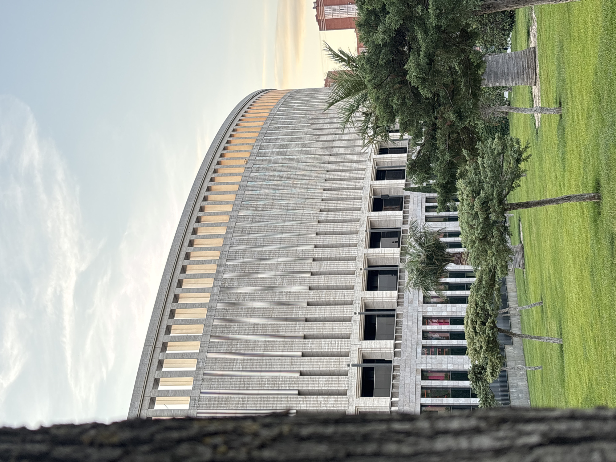 The image captures a large, curved building with a facade of vertical fluted panels and horizontal rectangular openings. The upper section of the building features a series of tall, narrow windows or recesses with a golden hue, suggesting sunlight. Below, there are rows of darker, rectangular windows. The building is set against a pale sky with wispy clouds, indicative of either early morning or late afternoon.
In the foreground, a well-maintained green lawn is dotted with several small, sculpted trees with slender trunks. To the right, a palm tree and other dense green foliage are visible, partially obscuring the building. In the distance, another brick building with visible windows can be seen. A portion of a large tree trunk occupies the left side of the frame, creating a foreground element.
This building is identified as the Assembly of the Legislative Body of Krasnodar Krai, located in Krasnodar, Russia.
