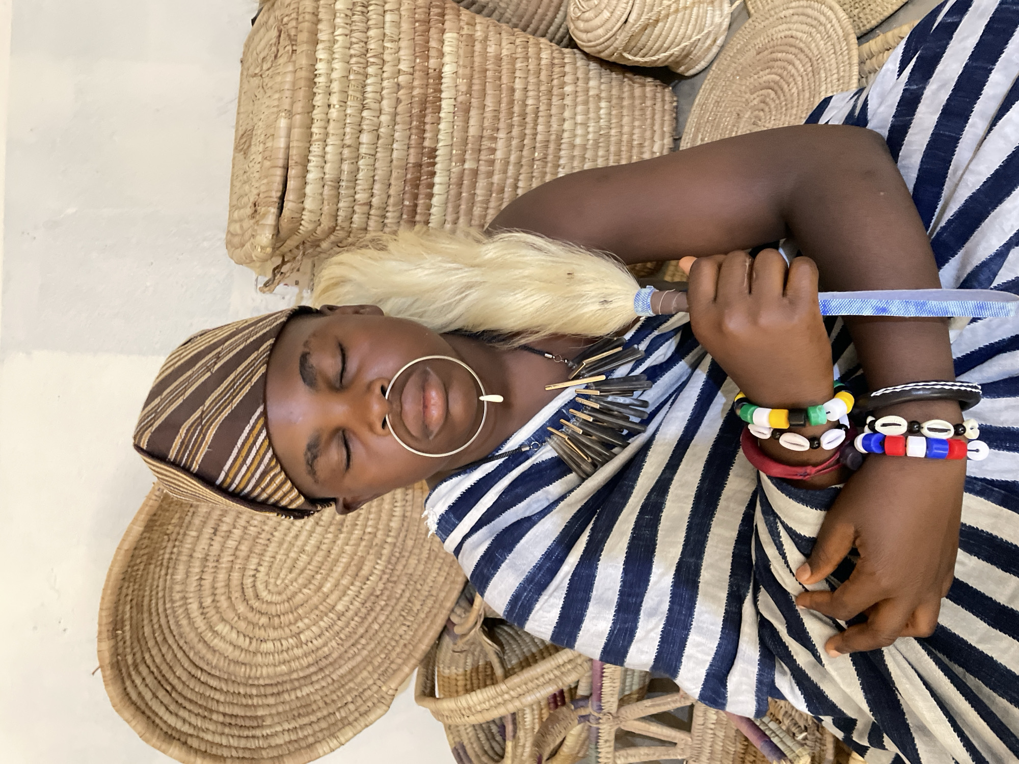 A person with closed eyes, wearing a brown-striped head covering, a large ring through the nose, a necklace of irregular brown pieces, and a blue and white striped garment, is holding a blue and brown handled object in their right hand. The person also wears multiple colorful bracelets on the right wrist. A pale blonde headdress or wig piece rests near the back of the person's head. The person is surrounded by various woven baskets and mats of different sizes and shapes. The location, Jos, Nigeria, is inferred from cultural objects in the image.
