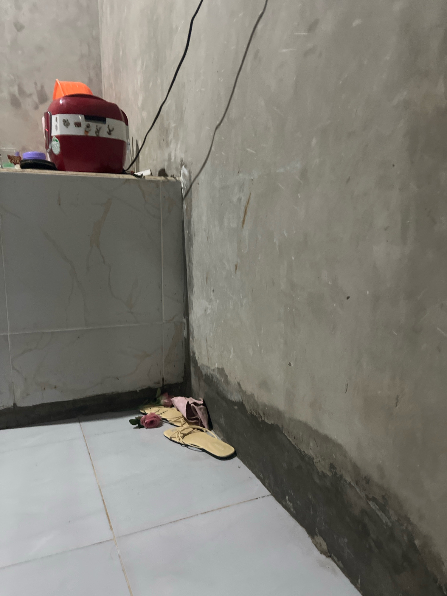 The image depicts a dimly lit, cramped interior space, likely a bathroom or kitchen corner. A red rice cooker with white trim and animal decals sits on a tiled surface, next to a purple object. A black cable extends upwards from the vicinity of the rice cooker. The walls are unfinished concrete, showing signs of moisture and discoloration near the floor. On the tiled floor, a pair of light yellow flip-flops and a pink garment are casually placed. A single pink rose lies near the flip-flops. The overall impression is one of a utilitarian, possibly modest living space. The location is identified as Kebumen, Indonesia.