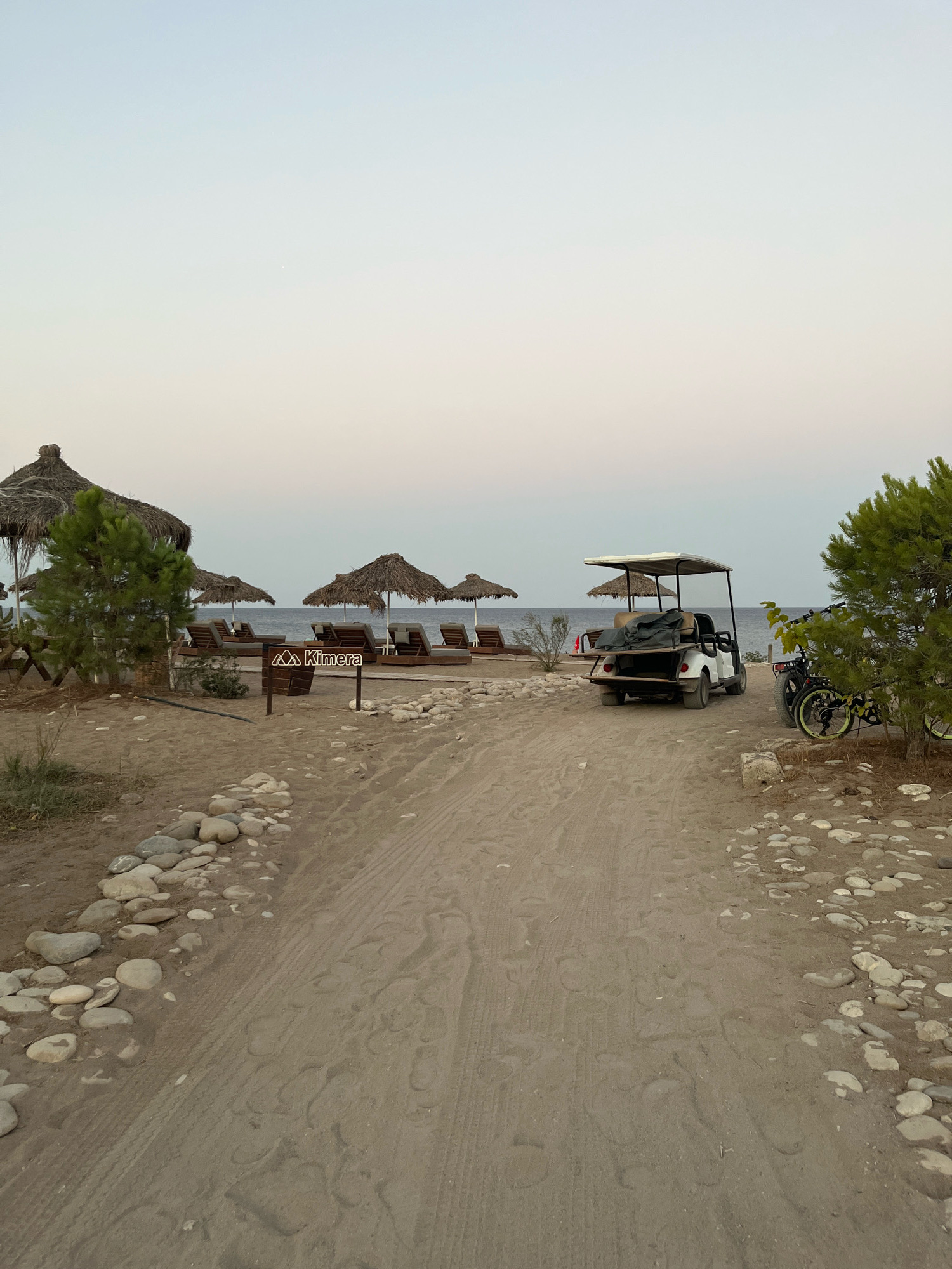 A sandy path leads towards the sea on a beach in Kemer, Turkey. On the left, several thatched umbrellas shade lounge chairs. A sign reading "Kimera" is visible next to the umbrellas. A white golf cart is parked on the right side of the path, with a bicycle behind it. The sea is calm and the sky is clear.