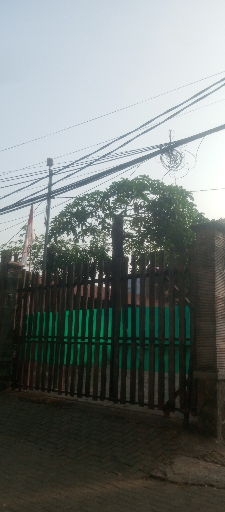 A wooden gate with green panels stands in front of a building. Above the gate, a flag is visible, as well as a light pole and numerous electrical wires. The sky is hazy, and the ground in front of the gate is paved with interlocking bricks. The image appears to have been taken in Kediri City, Indonesia.
