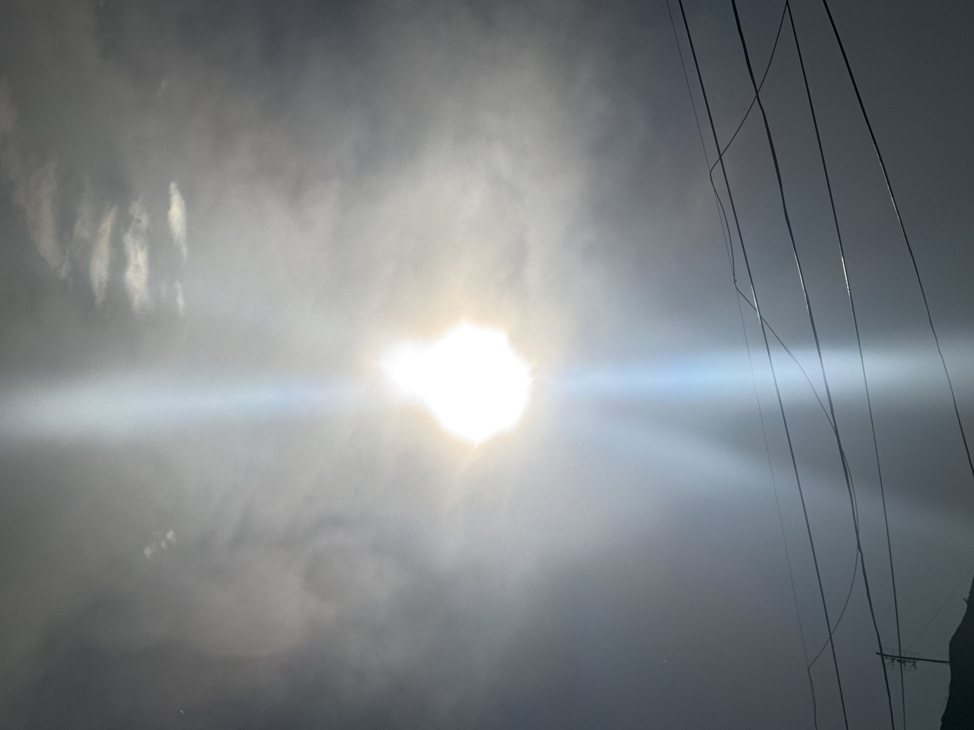The image shows a bright, hazy sun partially obscured by clouds. A strong beam of light emanates from the sun, creating a lens flare effect. In the foreground, several power lines are visible, stretching across the frame against the cloudy sky. The scene appears to be outdoors, with a hint of land visible in the lower left corner, suggesting a potentially elevated viewpoint. The overall impression is one of a bright, overcast day. The location is identified as Abovyan, Armenia.