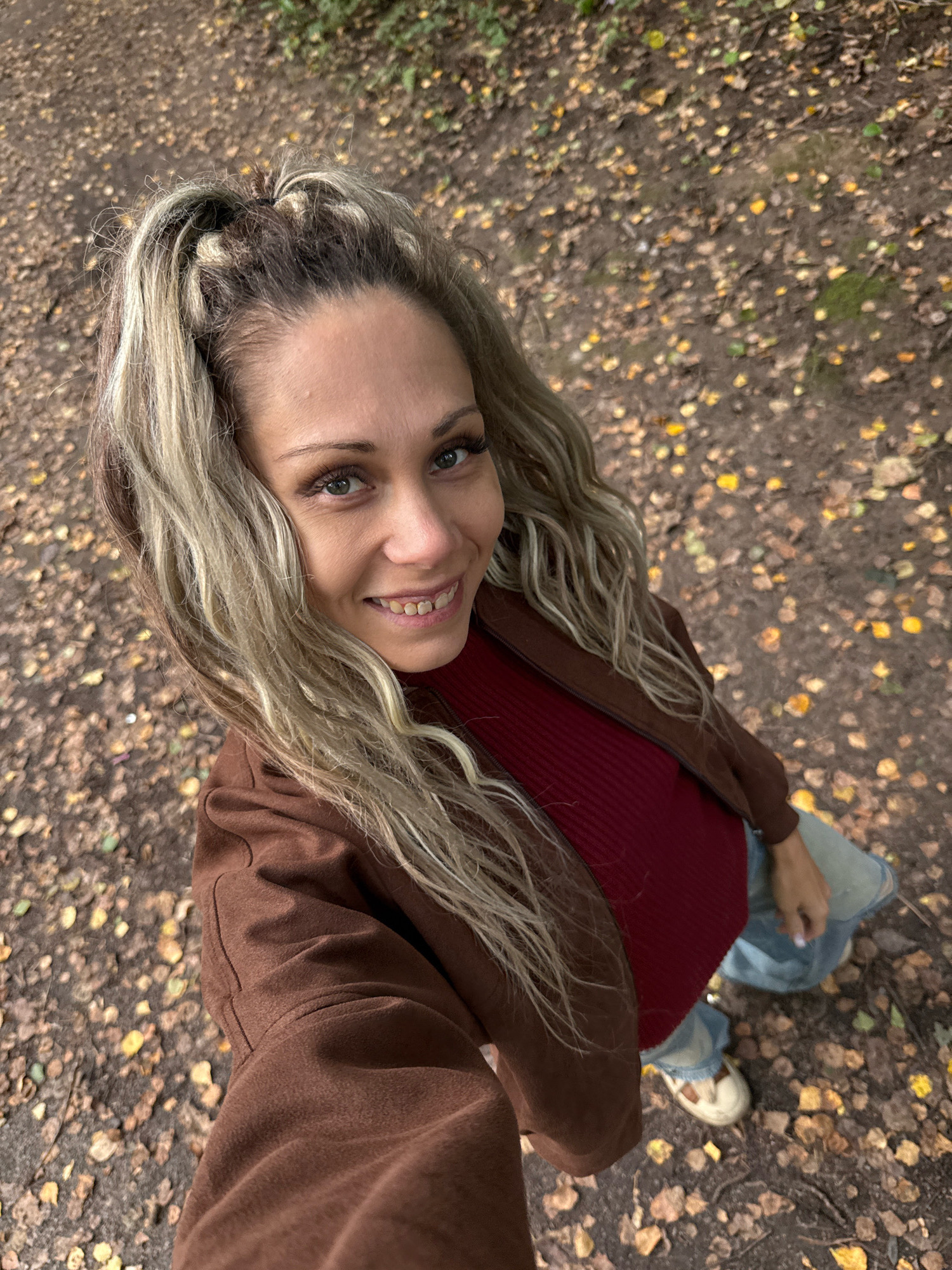 A high-angle, full-body selfie features a woman with blonde, wavy hair tied into two high ponytails. She is wearing a maroon ribbed top, a brown jacket, and light-wash jeans. She has on white and tan shoes. The woman is smiling and looking up at the camera, with her face slightly to the left. Her eyes are green, and she has on long false eyelashes and makeup. The background is a dirt path covered with fallen autumn leaves. The scene is likely outdoors, potentially in a park or wooded area, in the city of Dumino, Russia.