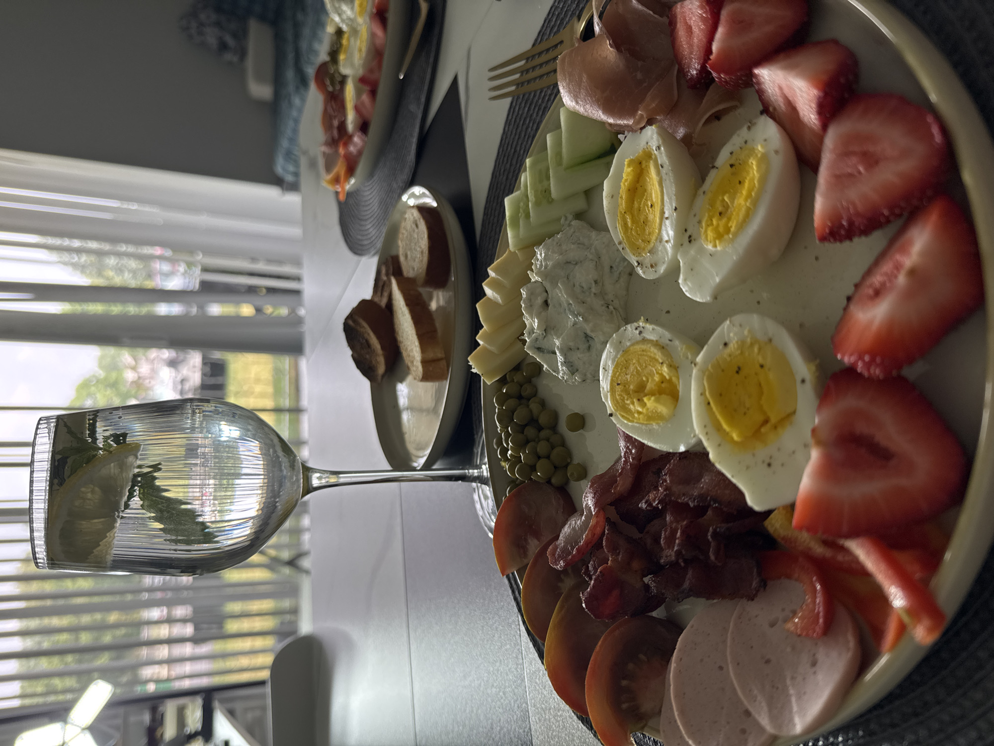 A close-up, eye-level shot captures a charcuterie board filled with breakfast and lunch items, set on a table in what appears to be a dining room. The board includes sliced hard-boiled eggs, bacon, sliced tomatoes, sliced ham, sliced red bell peppers, sliced strawberries, cucumber slices, a dollop of cream cheese dip, peas, and sliced bread on a separate plate. A tall glass of water with lemon and mint is positioned to the left of the charcuterie board. In the background, a window with sheer curtains reveals a blurred outdoor scene with trees. The scene is well-lit, suggesting it is daytime. The specific location within Wheeling, United States of America cannot be determined from the visual information provided.
