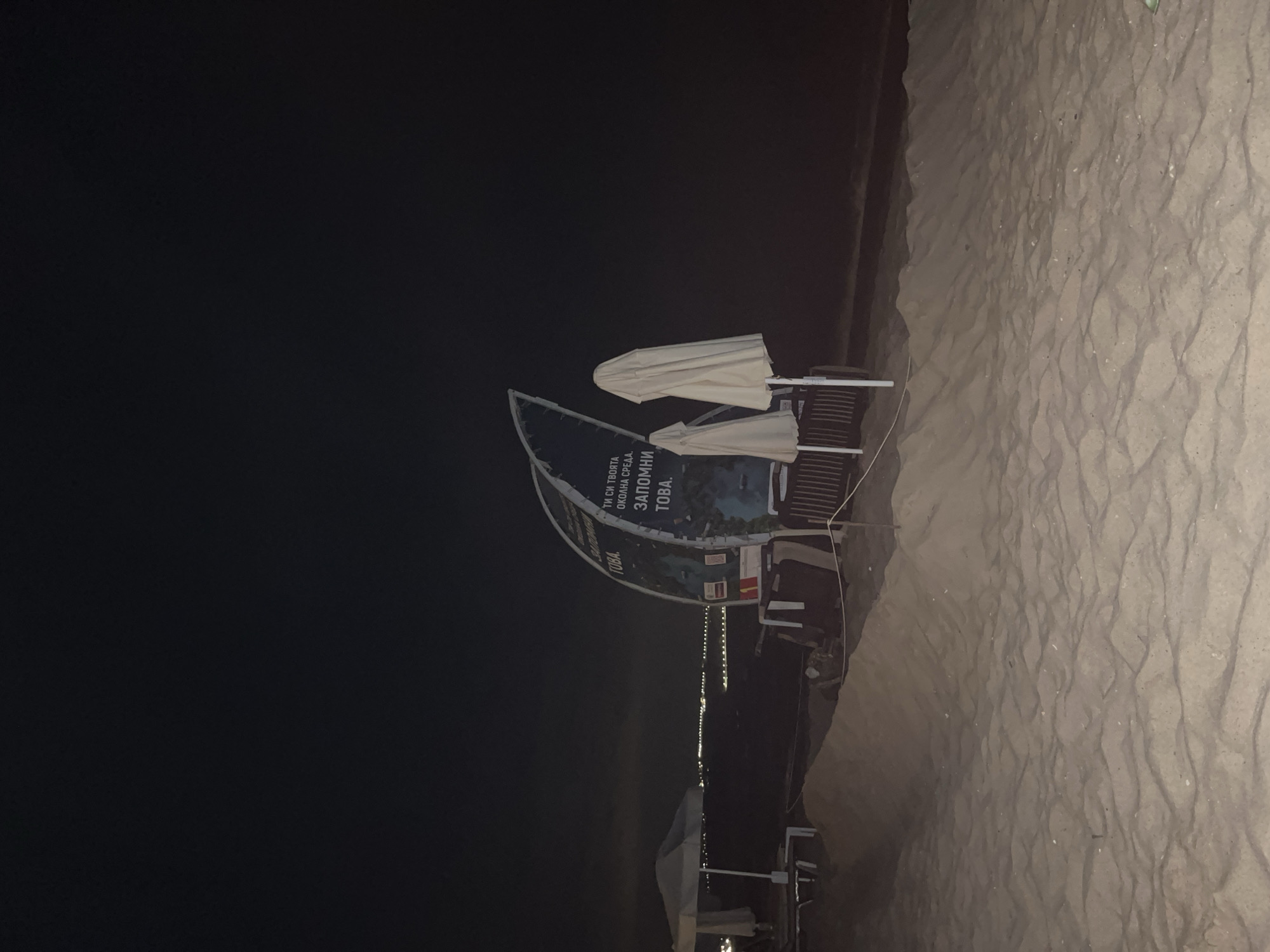 The image depicts a beach scene at night in Obzor, Bulgaria. In the foreground, there are sand dunes and rippled sand. In the midground, a large, curved billboard with text is visible, along with two closed beach umbrellas and parts of what appear to be beach chairs or lounge areas. A white beach umbrella is also visible to the left. In the background, a distant city skyline with lights is visible across the water. The overall atmosphere is dark and serene, characteristic of a beach at night. The text on the billboard is in Bulgarian and appears to be promotional or informational.