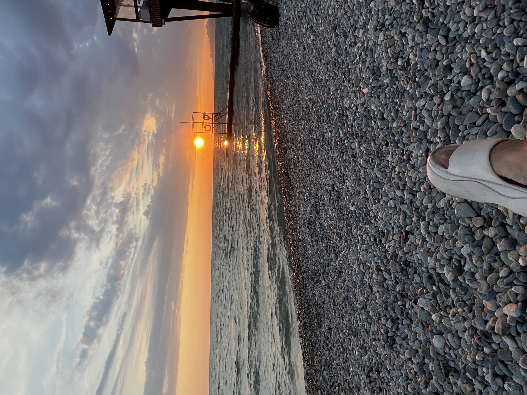A person's foot in a white sandal is visible in the foreground, resting on a pebble beach. The beach leads to the sea, where waves are gently rolling towards the shore. In the distance, the sun is setting over the water, casting a warm orange glow across the sky and reflecting on the sea's surface. A small pier with a lifeguard tower extends into the water. To the right, a wooden structure, possibly a part of a cafe or observation deck, stands on stilts over the water. The sky is a mix of grey and orange clouds, indicating the transition from day to night. This scene appears to be located on a beach in Sochi, Russia, given the architectural style of the lifeguard tower and the characteristic pebble beaches common in the region.