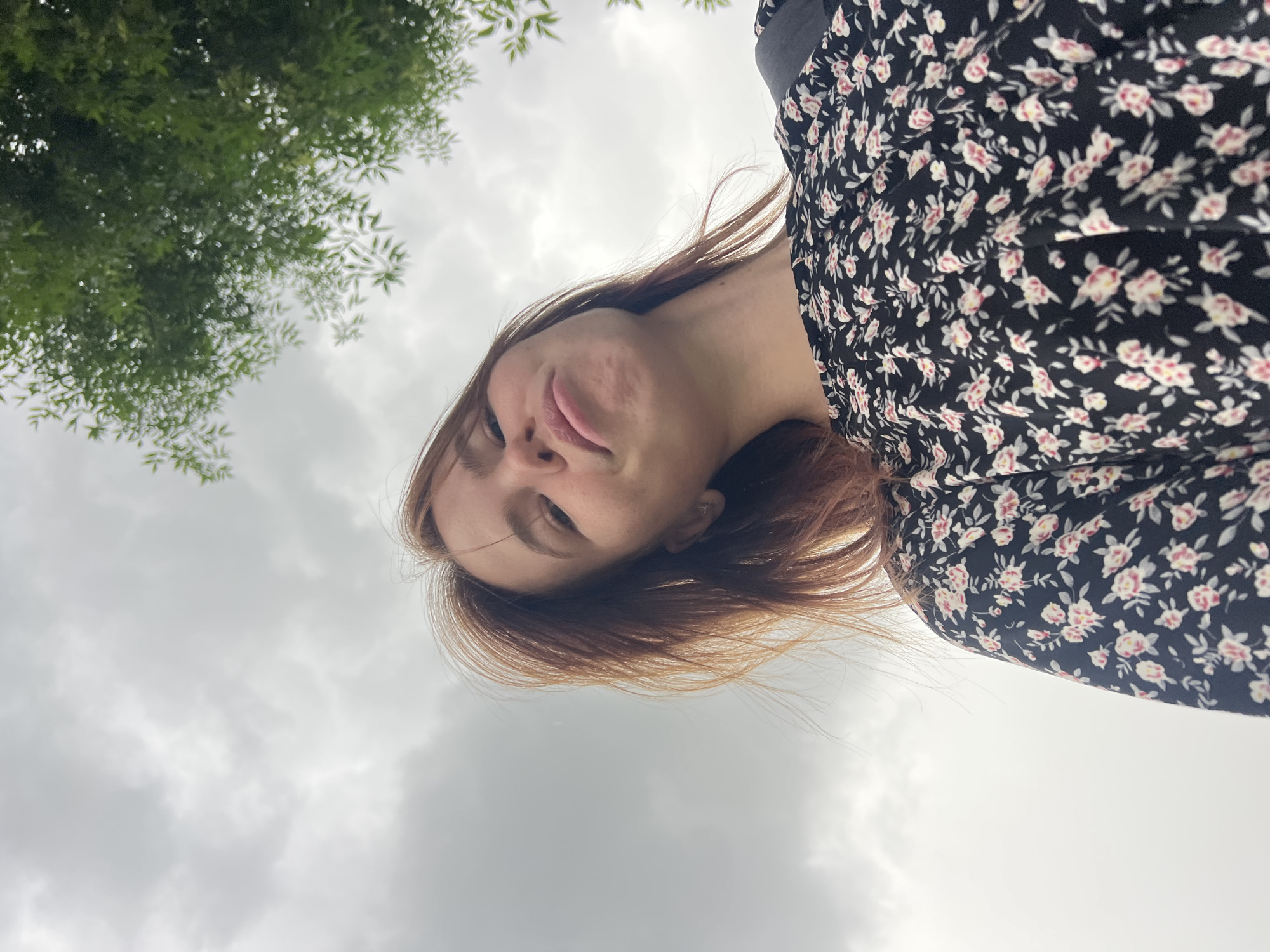 A low-angle, medium shot captures a young woman with shoulder-length, reddish-brown hair from the chest up, looking upwards towards the camera with a pout. She is wearing a black dress with a floral pattern of pink and white flowers. Her hair is windswept, and she has freckles across her nose and cheeks. A strap from a backpack is visible over her right shoulder. The background consists of a cloudy, overcast sky and the blurred green leaves of a tree. The scene appears to be outdoors in a park or natural setting. Given the prompt's specification, this photo was taken in Chaikovsky, Russia.