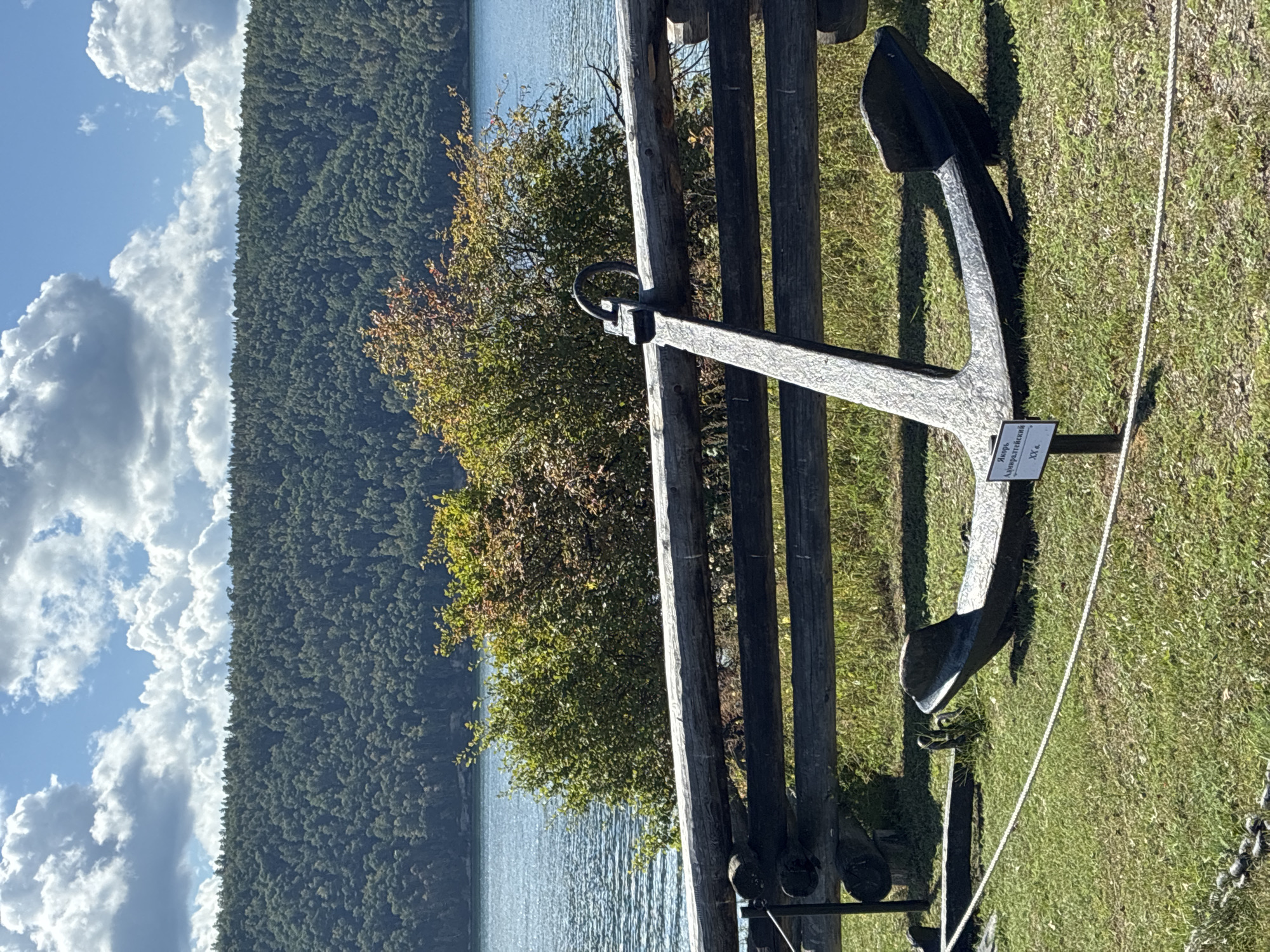 The image depicts an outdoor scene with a large, black metal anchor prominently displayed in the foreground. The anchor rests on a grassy area, partially in the shade cast by a rustic wooden fence constructed from logs. Behind the fence, a calm body of water, likely a river or lake, is visible, with a dense, tree-covered hillside forming the backdrop. The sky above is a bright blue with scattered white clouds. A small white sign with Russian text is affixed to a pole near the anchor. The location is identified as the Taltsy Museum in Irkutsk, Russia, an architectural-ethnographic museum.