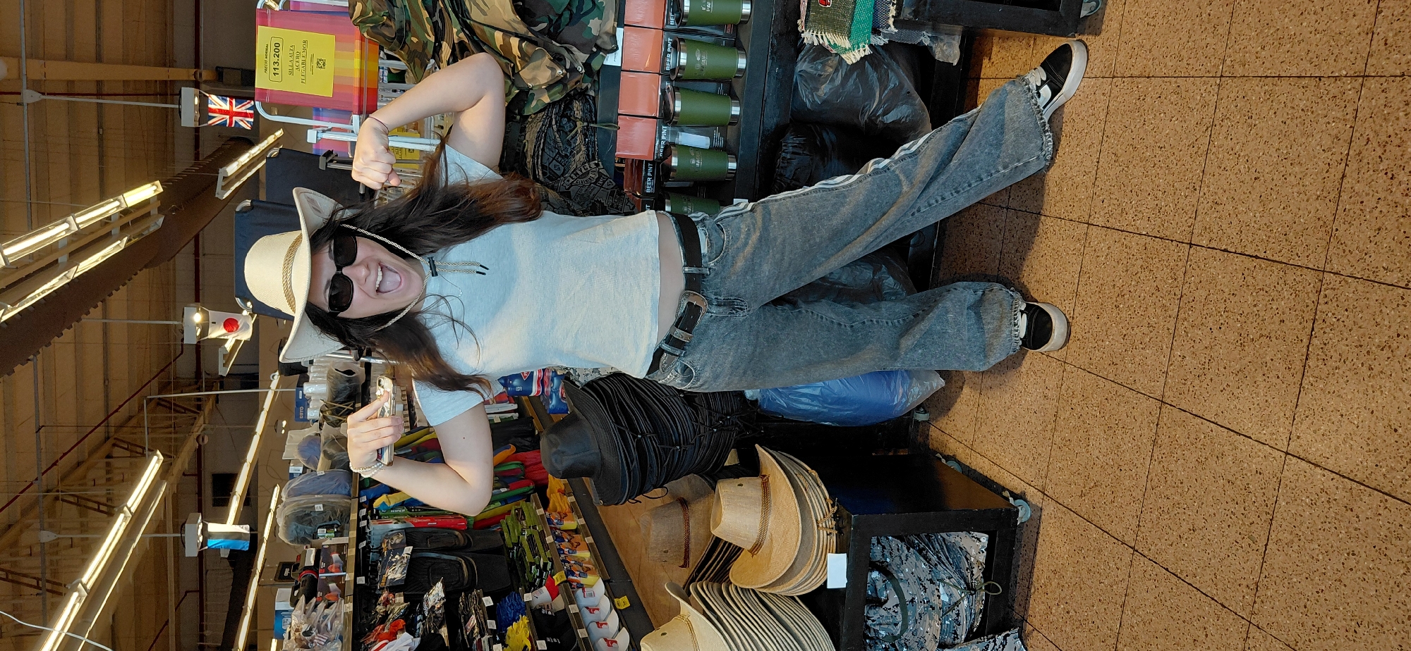 A young woman wearing a light blue t-shirt, acid-washed baggy jeans, sunglasses, and a white cowboy hat with a tan band poses assertively in an aisle of a retail store. Her arms are flexed, showcasing her biceps, and her mouth is open in an excited expression. The store appears to be selling outdoor and camping equipment, with shelves of merchandise visible in the background, including stacks of hats, camouflage gear, and green tumblers. The flooring is tiled with a speckled pattern, and overhead fluorescent lights illuminate the scene. The woman is standing in front of a display of various hats, including a large stack of black ones. A blue plastic bag is situated to her left. A yellow price tag with Spanish writing is visible on a folding chair in the background to the right. The environment suggests a commercial establishment, possibly a department store or a specialty retailer, located in Encarnación, Paraguay, indicated by the presence of flags from Japan and the United Kingdom, as well as a Spanish language price tag.