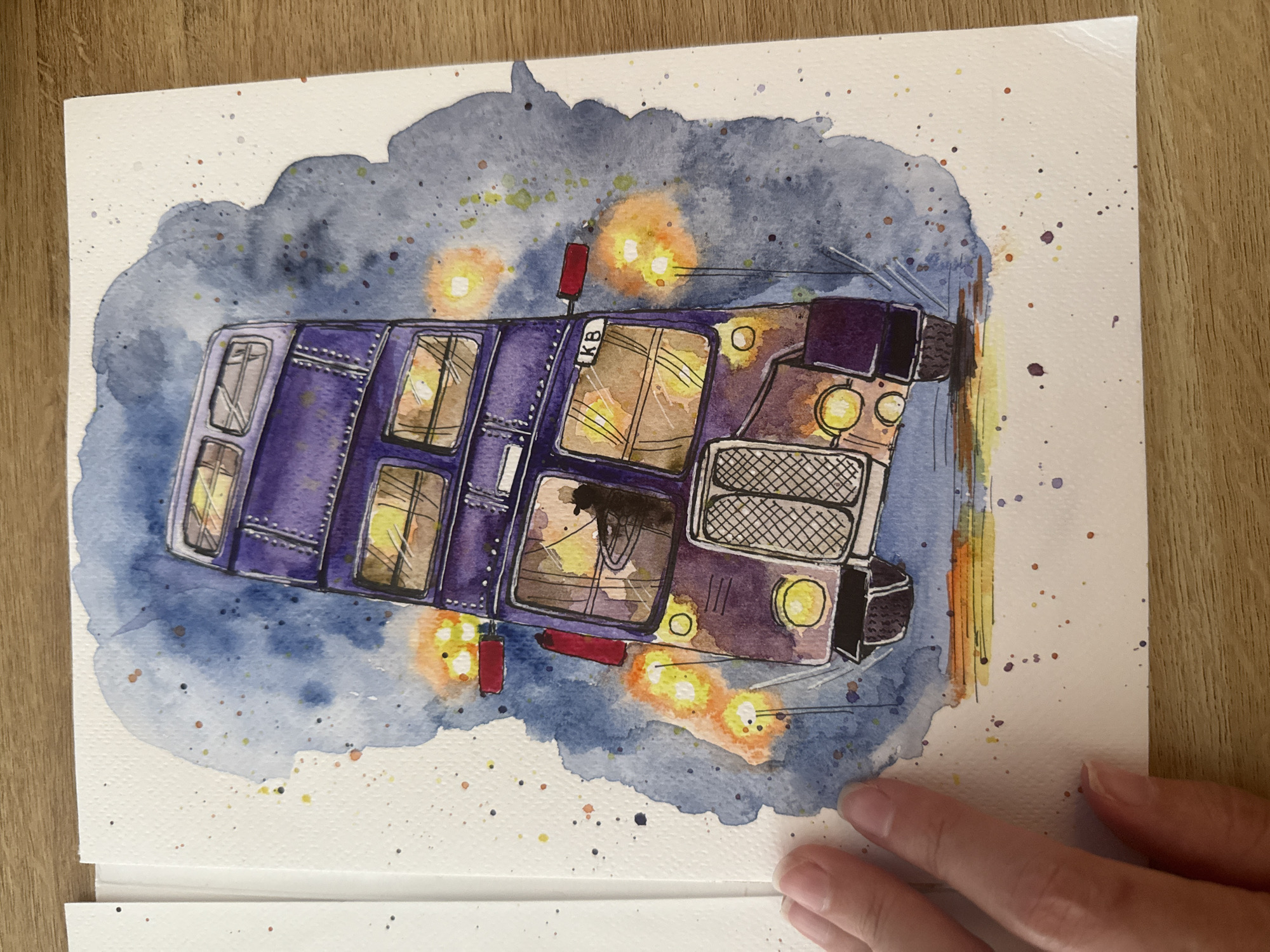 The image shows a watercolor painting of the Knight Bus, a purple double-decker bus from the Harry Potter series. The bus is depicted in motion, with its windows lit and steam or smoke effects around it. The painting is on textured paper, and the background is a wash of blue and orange watercolor with splatters of various colors. A human hand is visible on the left side of the image, touching the edge of the paper. The scene is not identifiable as being in Wrocław, Poland, or any specific city.