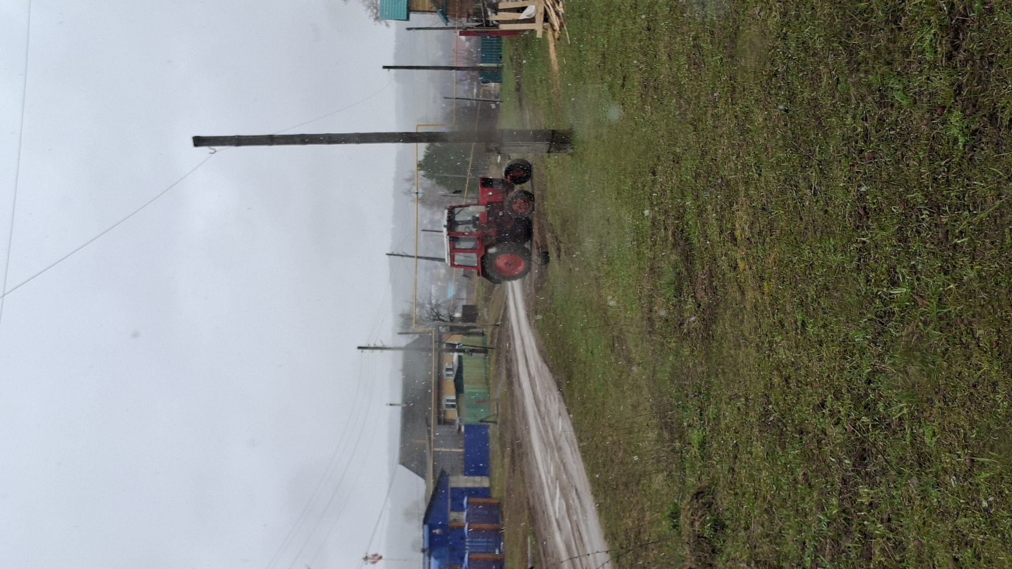 The image depicts a rural scene in what appears to be a village in Большой Букор, Russia. A muddy road cuts through the center, flanked on one side by a grassy field and on the other by a collection of houses and buildings. A red tractor is parked on the road near a utility pole. Several other utility poles with wires are visible in the background, running along the road. The sky is overcast, and what appears to be either rain or snow is falling. The houses are simple structures, some painted in bright colors like blue and green. There are no people visible in the image.