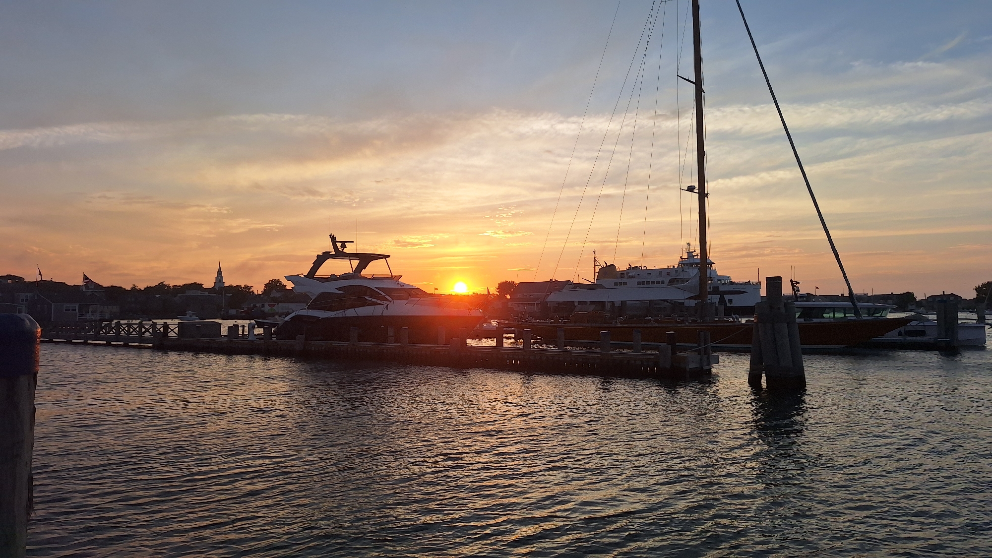 This is a wide-angle, eye-level photograph of a harbor at sunset on Nantucket Island, Massachusetts, USA. The sun, a brilliant orange orb, is low on the horizon, casting a warm glow across the sky and water. The sky is a gradient of blues, oranges, and yellows, with streaks of thin clouds adding texture.
Several boats are docked in the harbor. In the foreground, a large white yacht with a dark hardtop is prominently featured, its sleek lines silhouetted against the sunset. Next to it, a tall sailboat mast with rigging stretches towards the sky. In the background, a larger white ferry-like vessel is docked, and further in the distance, a small church spire can be seen rising above the rooftops of buildings that line the shore.
The water in the harbor is calm, with gentle ripples reflecting the colors of the sunset. Wooden pilings and parts of a pier are visible, further anchoring the scene to the waterfront. The overall mood is serene and picturesque, capturing the beauty of a coastal sunset. There are no people visible in the image.