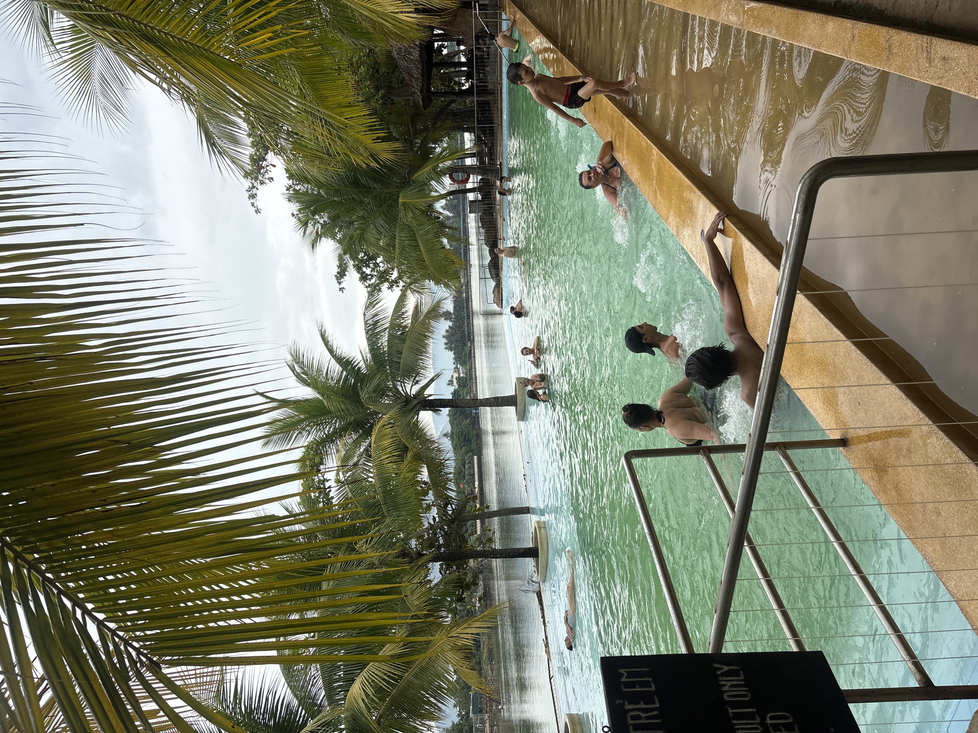 The image depicts an outdoor infinity pool with people swimming and relaxing. Palm trees frame the scene, with a body of water in the background that appears to be a river or the sea. In the foreground, a black sign with white text is partially visible, with "TRE EM" and "ULT ONLY ED" legible. Several individuals are in the pool, some are splashing, others are floating or sitting on the edge. A young boy in swim trunks is standing on the pool's edge. The water is a vibrant green, and the pool deck is a warm, sandy color. The sky is overcast. This appears to be the infinity pool at Vinpearl Land Water Park in Nha Trang, Vietnam, a popular tourist destination known for its amusement park and water park attractions.
