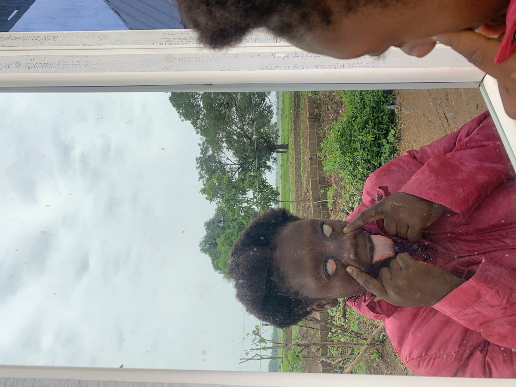 A photograph taken in Agban, Nigeria, shows a dark-skinned individual in a red jacket positioned at an angle in front of a mirror, pulling their eyes open with their fingers and sticking out their tongue. The reflection reveals the individual and a background featuring a grassy area with a large tree, possibly indicating a rural or outdoor setting. The sky is overcast. The mirror has white framing with some writing along the frame.