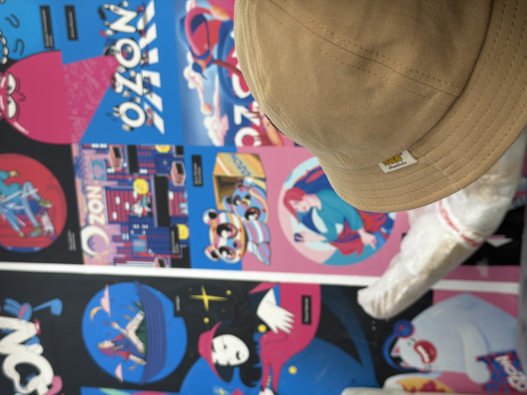 A beige cap with a visible tag is positioned in front of a wall covered with colorful, abstract posters. The posters are arranged in rows and feature a variety of designs, including cartoon-like characters, cityscapes, and portraits. The scene is located in Chaikovsky, Russia.