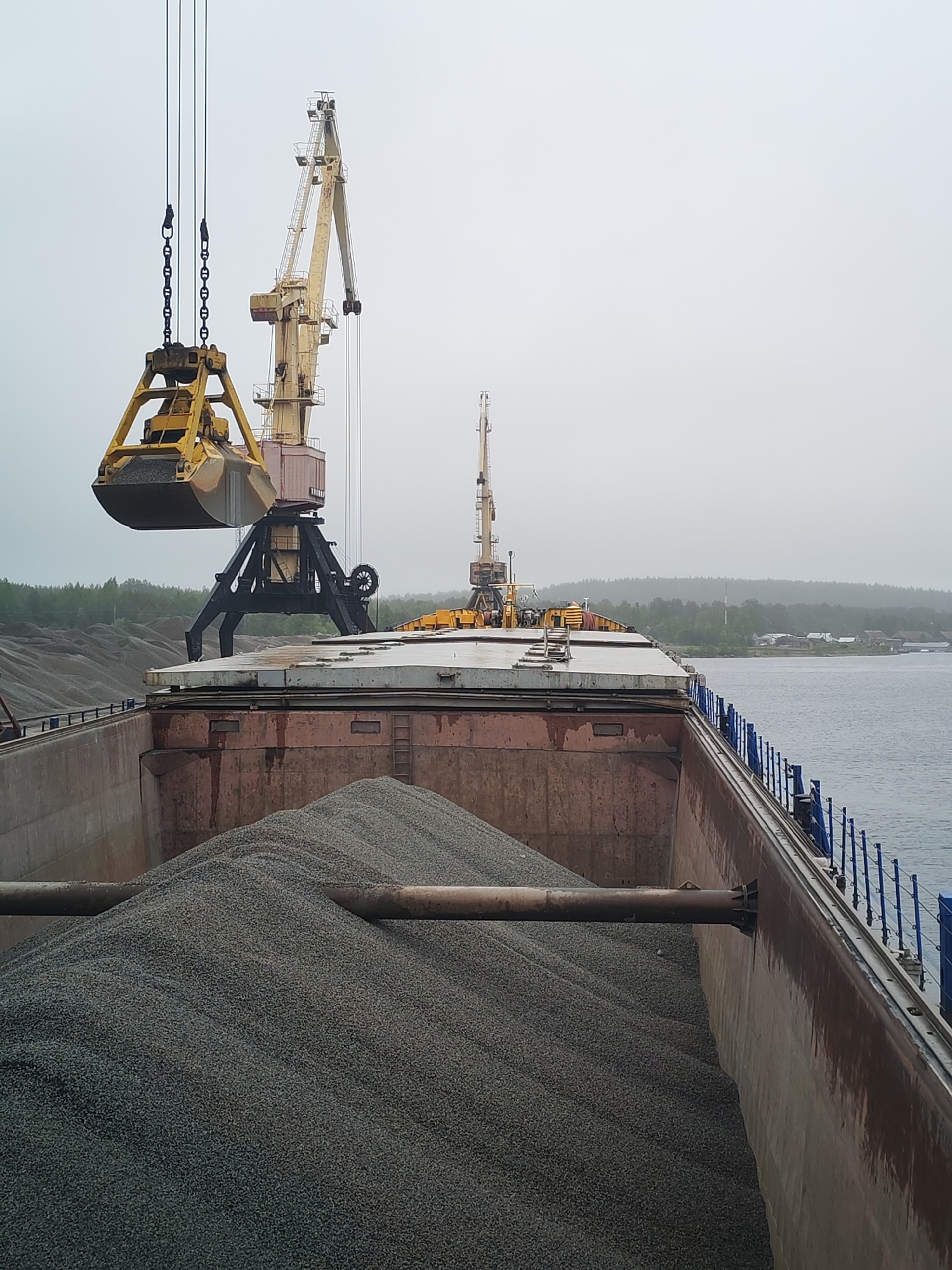 A cargo ship is being loaded with gravel by a crane in Shalsky, Russia. The ship is filled with a large mound of gray gravel, and a yellow crane is suspended over the hold, its grabber poised to release another load. In the background, a second crane is visible on the deck of the ship. Beyond the ship, the shoreline of Shalsky is visible, with trees and buildings lining the coast. The sky is overcast and gray.