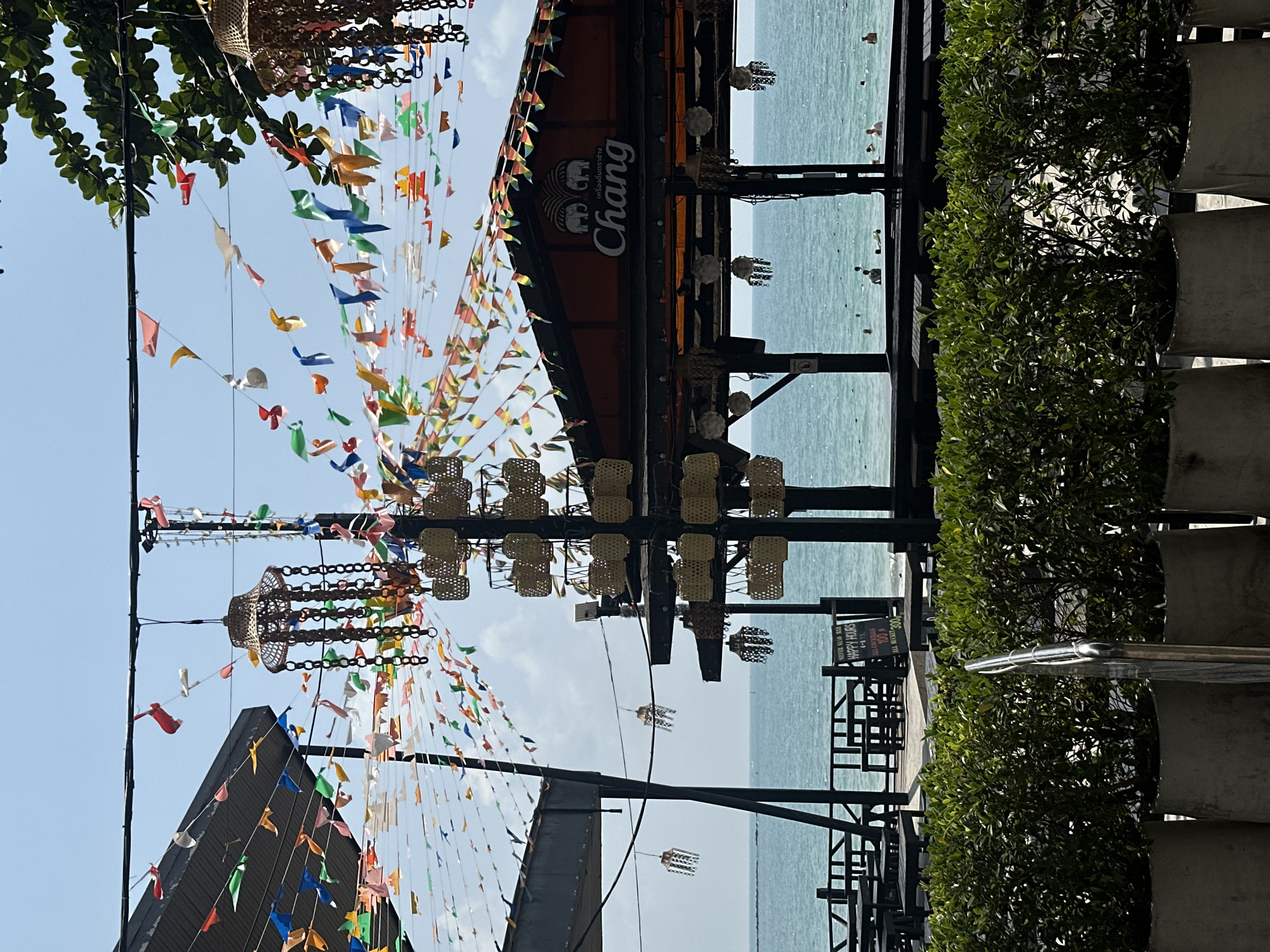The image depicts a coastal scene in Ao Noi Na, Thailand. It features a structure with a "Chang" beer sign, positioned near the sea. Decorative elements such as colorful flags and woven lanterns are suspended above the walkway. A row of potted plants line the right side of the image, and there is a sign advertising "Happy Hours" at a nearby bar. The beach and sea are visible in the background.