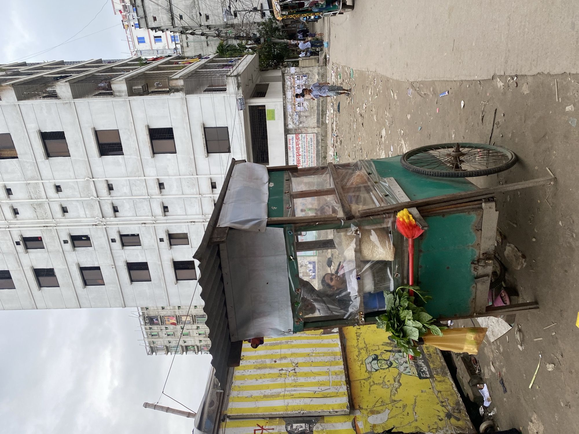 In Dhaka, Bangladesh, a street scene unfolds with a food cart as the central subject. The cart, painted green with a corrugated metal roof and clear plastic sheeting, displays an assortment of goods. Inside, a person is visible behind the counter. To the left of the cart, a yellow container with vertical white stripes is adorned with a graphic of a chef and text. The ground is unpaved and strewn with debris, indicative of a bustling urban environment. In the background, tall, multi-story buildings rise against an overcast sky. A young boy walks along the dirt path in the middle ground. Further in the distance, a rickshaw is visible, adding to the sense of urban activity. The overall impression is one of everyday life in a densely populated city.