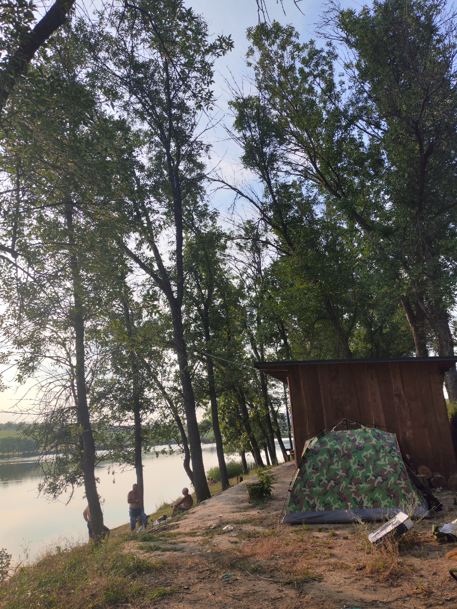 The image depicts a campsite on the bank of a body of water, likely the Nistru River, near Găleștii Noi, Moldova. Tall trees with green foliage dominate the background, with their branches reaching towards a partly cloudy sky. In the foreground, a camouflage tent is set up on the sandy ground, adjacent to a small wooden structure. Three men are present near the water's edge. One man, wearing blue jeans, stands near a tree. Another man, shirtless, is also standing, facing away from the camera. A third man, wearing brown shorts and a light-colored shirt, is seated on the ground, facing the water. The overall scene suggests a relaxed outdoor setting, possibly for fishing or recreation. The presence of trees, water, and a campsite points to a natural, undeveloped area.