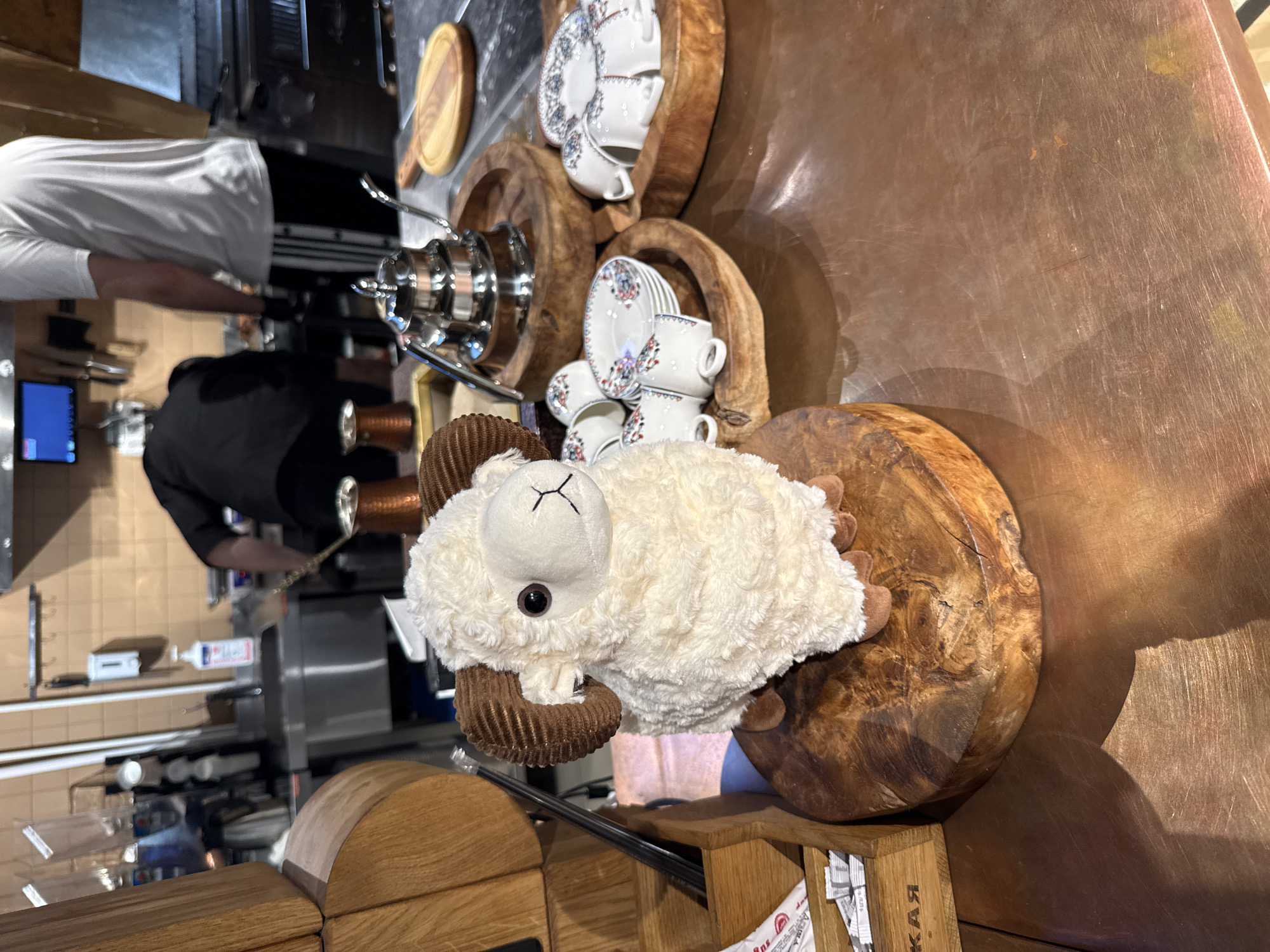 The image shows a blurred interior scene. In the background, there is a glimpse of people in what appears to be a kitchen or food preparation area. The foreground features a plush, white ram toy placed on a wooden surface, likely a countertop. A serving or tea set is also present. The photograph seems to have been taken in a restaurant or similar establishment, potentially in the city of Khimki, Russia, based on the presence of the Russian text on the box.