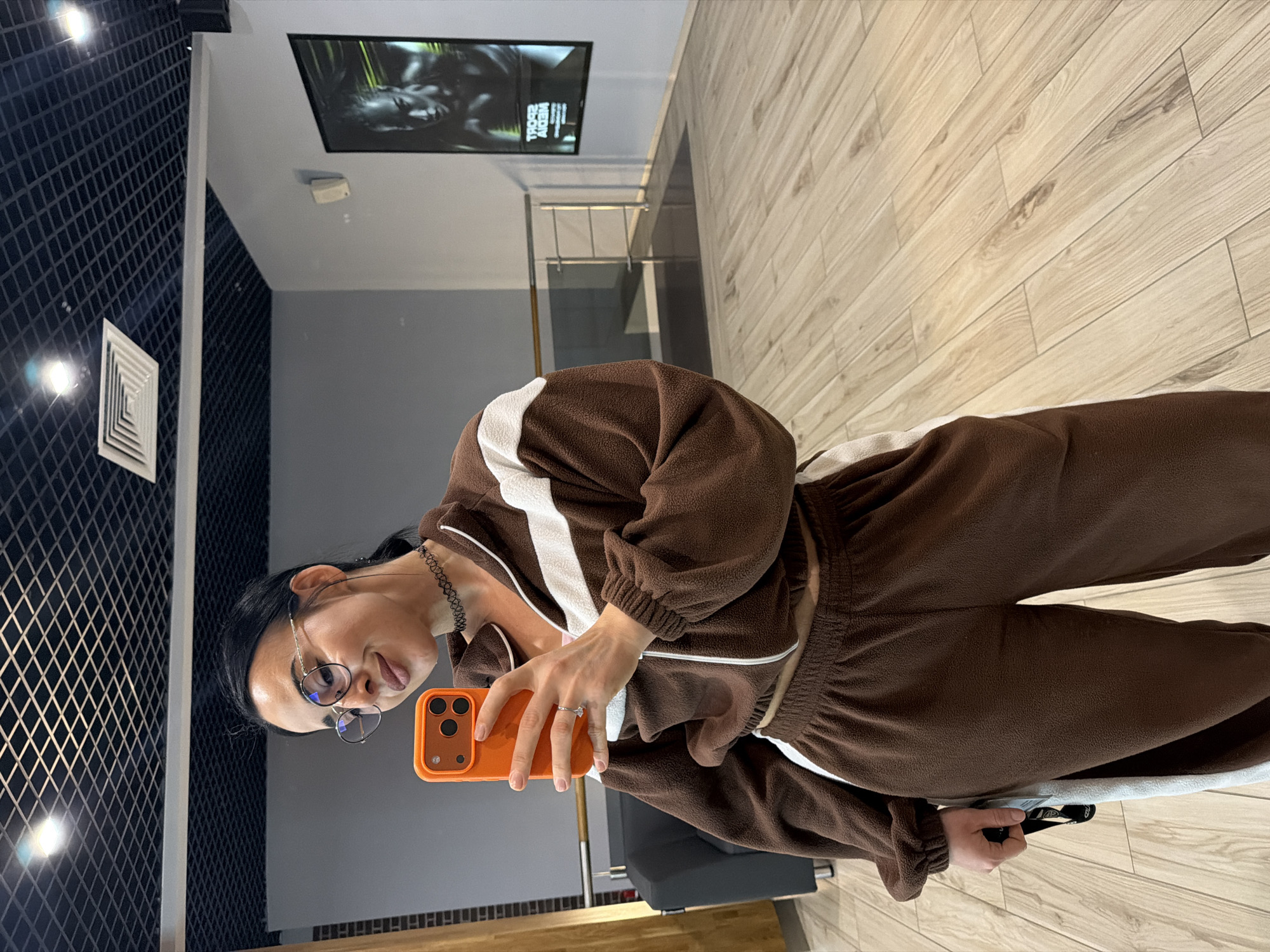 A young woman is captured taking a mirror selfie in a modern indoor setting, located in Saint Petersburg, Russia. She stands slightly to the left of the frame, holding an orange smartphone, likely an iPhone, to capture her reflection. She has dark hair, wears round-rimmed glasses, and a black choker necklace. Her expression is neutral, focused on the phone. She is dressed in a comfortable-looking brown tracksuit with white stripes, made of a fleece-like material. A ring is visible on her right hand.
The background reveals a contemporary interior. To her left, a dark wall features a grid pattern, reflecting a bright light source. The floor is covered in light-colored wooden or wood-effect laminate. The ceiling is light-toned, fitted with a white square ventilation grate and recessed lights. In the upper background, a flat-screen television is mounted on a light gray wall, displaying an image that appears to be of an animal, possibly a gorilla or chimpanzee, alongside visible text. The text on the screen reads "TROPO ALIEN" with additional, smaller text below it that is difficult to decipher. A glass railing is also visible in the background, suggesting a multi-level space or a mezzanine. The overall environment appears well-lit, clean, and modern.