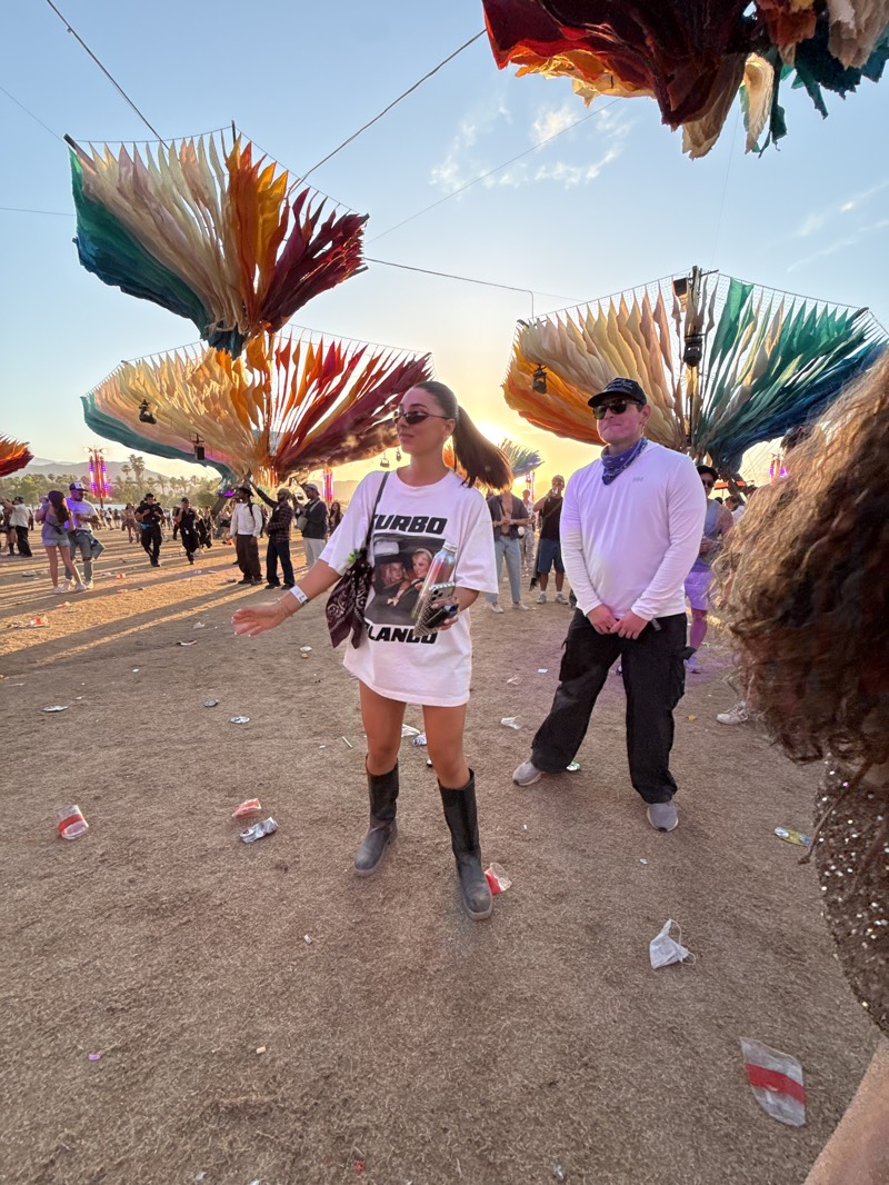 The photograph depicts a scene at what appears to be a music festival in Indio, United States, during daylight hours with the sun setting in the background. Several people are visible in the scene. A woman is standing prominently in the center, wearing a white t-shirt dress, black boots, and sunglasses, with her hair pulled back in a ponytail. She has a bag over her shoulder and appears to be gesturing with her right hand. To her right stands a man dressed in a white long-sleeved shirt, black pants, a baseball cap, and sunglasses, with a bandana around his neck.
In the background, there are numerous other individuals, some walking and others standing, suggesting a crowded environment. Overhead, there are colorful decorative structures resembling petals or fabric suspended from wires, adding to the festival atmosphere.
The ground is covered in dirt and littered with discarded cups and wrappers, indicating a lack of cleanliness in the event space. Overall, the image captures a moment at a lively, but somewhat messy, outdoor event.