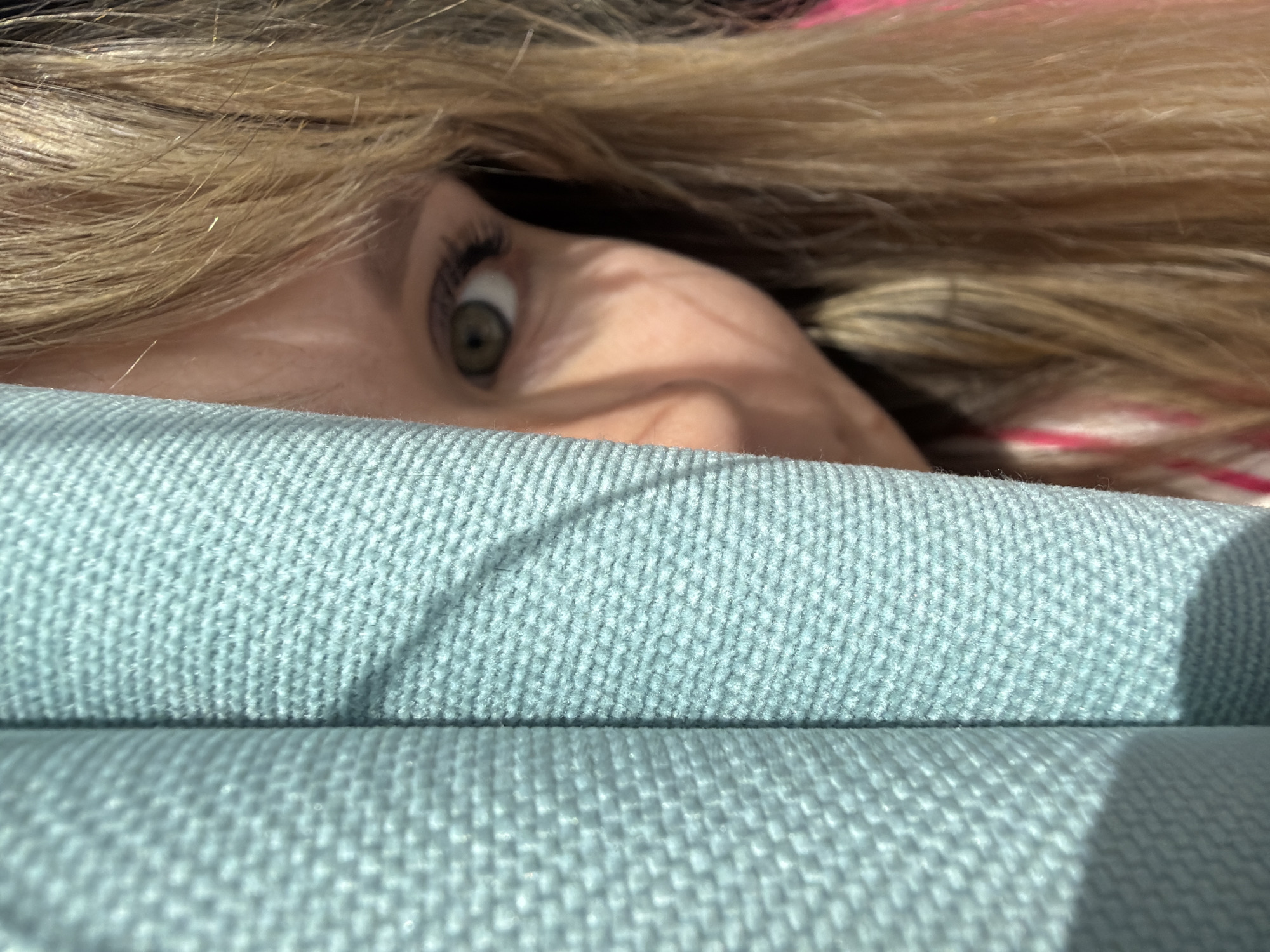 A close-up shot presents a person with blond hair peeking over the edge of a light blue, textured surface, possibly a couch or cushion. Only part of the face is visible, mainly the eye and the area around it. The eye is green, and the expression suggests surprise. The environment appears to be indoors, as indicated by the presence of furniture. There are no specific details that suggest the location to be Chaikovsky, Russia.